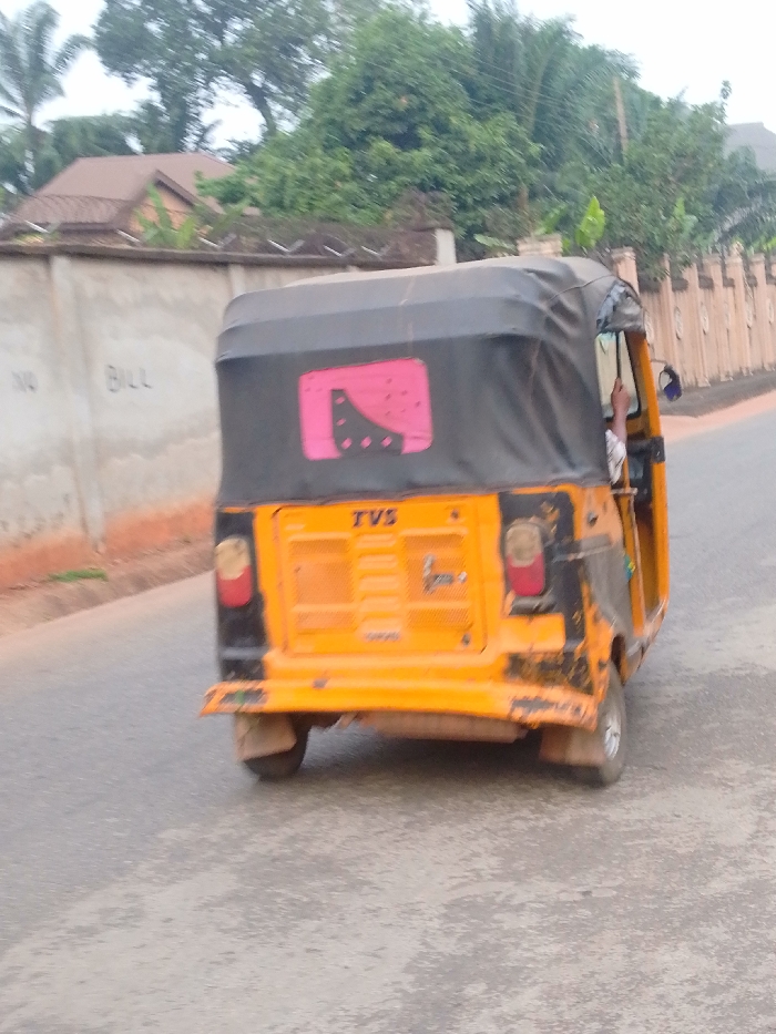 A three-wheeled yellow and black TVS auto-rickshaw, commonly known as a Keke Napep, is depicted from its rear, slightly angled right, in motion on a grey asphalt road in Alor, Nigeria. The vehicle features a black fabric soft top with a pink and black graphic sticker on the rear window. The "TVS" brand name is visible on its yellow rear panel. A person's right arm and a portion of a white shirt are visible inside the cabin on the right side. To the left, a light-colored concrete wall topped with razor wire fencing bears the graffiti "NO BILL." Behind this wall, dense green foliage and a brown-roofed building are partially visible. To the right, a brown decorative wall with pillar-like structures extends along the road, also backed by green vegetation.