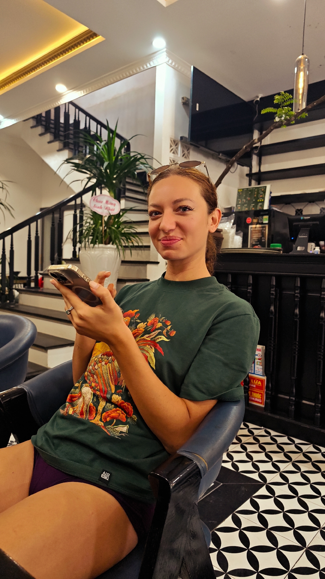 A young woman with light skin and dark hair tied back sits in a dark blue chair with wooden armrests, looking directly at the camera with a slight smile. She wears a dark green t-shirt with a colorful, ornate floral design and purple shorts. Sunglasses rest on top of her head. She holds a smartphone with a clear case in both hands, her left hand displaying a ring on her ring finger.
The setting appears to be an indoor establishment, possibly a cafe or lobby in Phố Ràng, Vietnam, given the overall decor and visible text. Behind her, a staircase with black banisters and light-colored steps ascends. A large potted plant in a white planter is situated near the staircase, next to a small pink sign. To the right of the woman is a dark-colored counter or bar area, adorned with various items and smaller, mostly illegible signs. A modern light fixture with a long bulb hangs from the ceiling in the background. The floor is covered in distinctive black and white geometric patterned tiles. The ceiling features decorative molding and gold-colored trim.
The woman's expression is engaging and cheerful. The indoor lighting suggests a well-lit environment, but the time of day is not explicitly visible.
Visible text includes "Chúc Mừng Sinh Nhật" on the pink sign (Vietnamese for "Happy Birthday"), and a small, illegible white tag with black text on the lower left hem of her t-shirt. Some faint text like "LUA" and "OKING" can be seen on a sign on the counter.