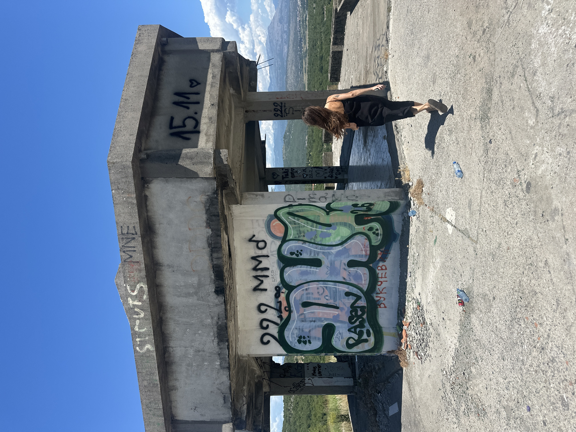 In Gornji Kokoti, Montenegro, an abandoned concrete structure, likely a former observation point or unfinished building, stands on a hill overlooking a valley. The structure is heavily graffitied. On the top horizontal concrete slab, the words "ST. CUTS MNE" are spray-painted in white, with "15.11" followed by a heart spray-painted in black on a lower section of concrete. On a vertical concrete wall, large graffiti in green and blue spray paint reads "SDH" with "RASEN" written below in smaller red letters, and "ВУКЧЕВ И" is written in red spray paint at the bottom. A woman with long brown hair, wearing a black dress and sandals, is walking away from the camera towards the right side of the frame, her shadow cast on the concrete ground. The ground is littered with debris, including broken concrete and several plastic water bottles. The background shows a lush green valley with distant mountains under a clear blue sky with scattered white clouds.