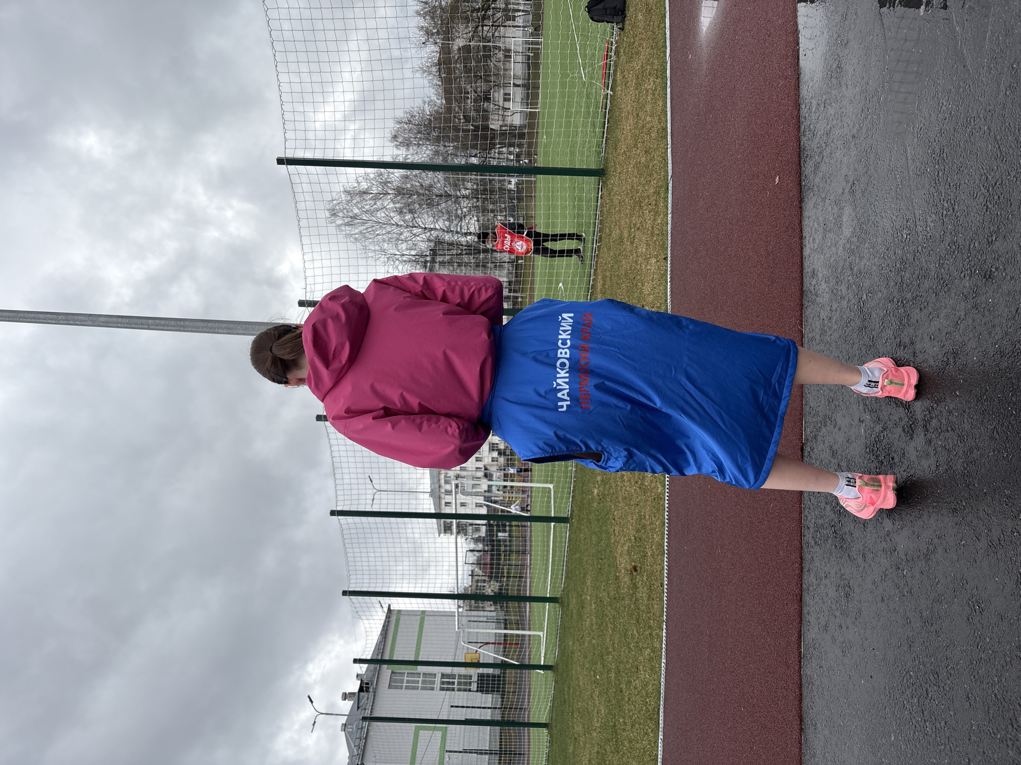 A vertical shot shows a person in a pink jacket and blue skirt, likely a referee or event staff member, standing on a wet, dark surface adjacent to a running track and a soccer field. The person is wearing pink running shoes. On the blue skirt are the words "Чайковский Пермский край" ("Chaikovsky Perm Krai"), which indicates that the location is in the city of Chaikovsky in the Perm Krai region of Russia. In the background, there is a green and white building, a soccer field with a net, and a person in a red garment, perhaps another referee. The sky is overcast. The photograph appears to have been taken in a sports complex or a park in Chaikovsky.