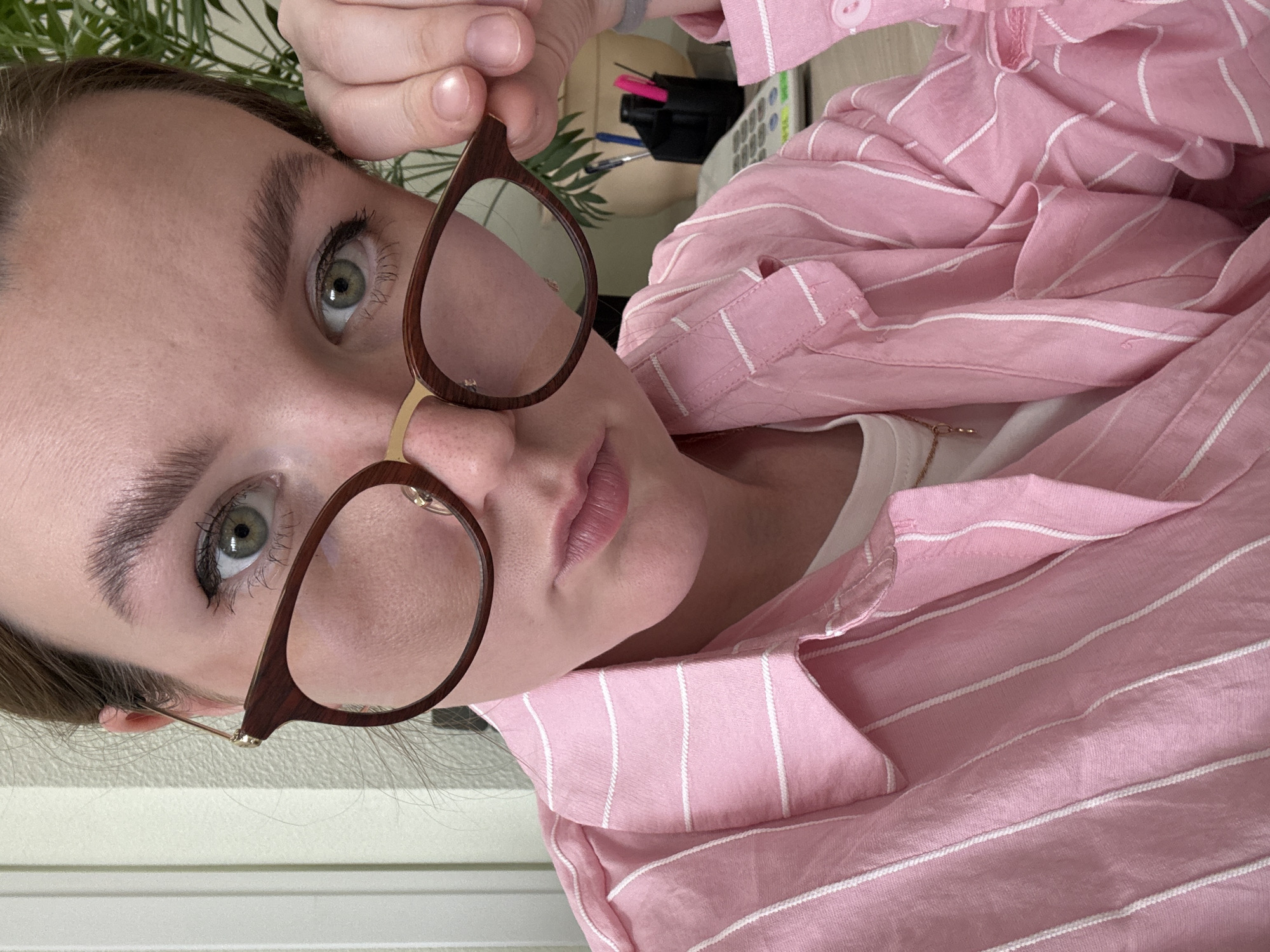 The media file is a close-up, high-angle, portrait-style photo of a young person wearing glasses and a pink and white striped shirt. The subject's face is angled towards the viewer, with their right eye and mouth clearly visible. They are wearing glasses with thin, light brown frames and appear to be looking at the camera. Their left hand is visible near their forehead, holding the top of the glasses. The background is blurred, but hints of a desk with office supplies, including what appears to be a calculator and a pen holder with pink and blue pens, can be seen. A leafy plant is also partially visible to the left. The lighting suggests it could be daytime, possibly indoors. The overall emotion conveyed is neutral or contemplative. There is no visible text in the image. The context provided indicates the location as Izhevsk, Russia.