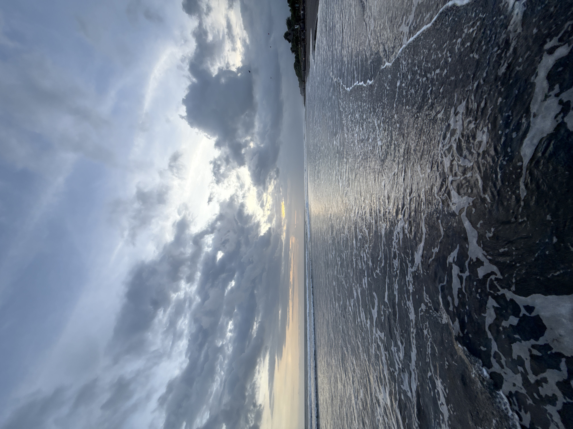 The image depicts a beach scene under a cloudy sky. The scene is divided roughly in half, with the sky on the left and the ocean on the right. The sky is filled with gray clouds, with patches of lighter blue visible. The sun is partially obscured by the clouds, creating a diffused light. The ocean is dark and turbulent, with white foamy waves crashing on the shore. The beach is barely visible, but there appears to be a line of trees or vegetation in the distance. The scene is located in Unknown, Unknown.