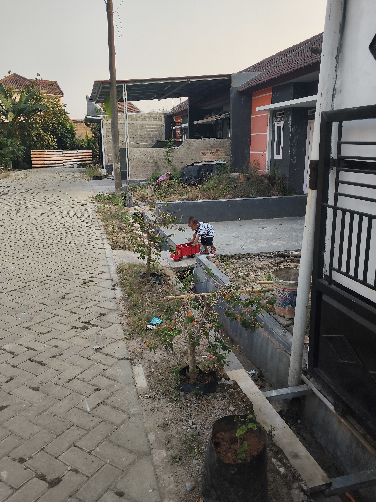 In Banjar Agung, Indonesia, a young child in a striped shirt and dark shorts plays with a red toy truck on a paved driveway. The child is near a concrete drainage ditch. To the left of the driveway, there are houses with tiled roofs, some under construction with exposed brickwork. Mature trees with large leaves, including banana trees, are visible near one of the houses. The area appears to be a residential street with several homes and a utility pole. In the foreground, a small plant in a black plastic bag sits on the edge of the driveway. The sky is overcast.