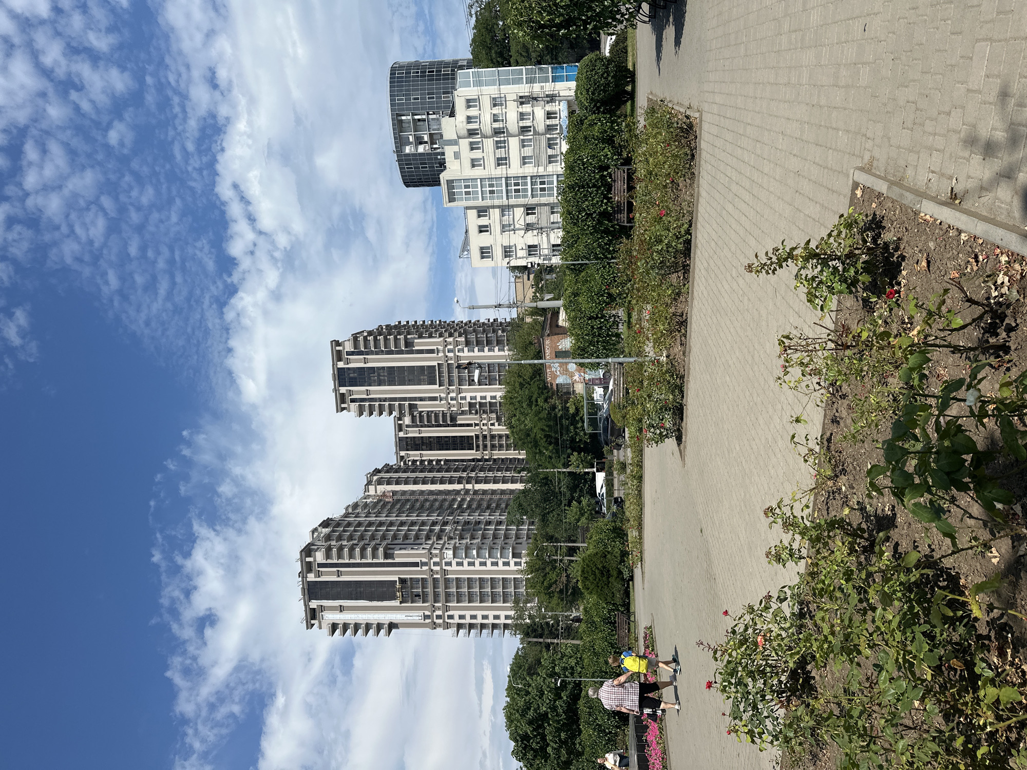 This is a daytime, vertical, eye-level shot of a public park area in Odesa, Ukraine. The foreground features flowerbeds with rose bushes. A paved walkway with a light gray, brick-like pattern runs through the center of the image, leading the eye toward the background. Two people, an adult and a child, are walking away from the camera on the walkway. The adult, on the left, wears a checkered shirt and dark shorts, carrying a black bag. The child, to the right, wears a bright yellow backpack and dark shorts. In the midground, there are more flowerbeds, benches, and trees lining the walkway. Beyond the park, several modern, high-rise buildings dominate the skyline, including a cluster of tall, beige apartment buildings and a distinctive cylindrical glass building. The sky is bright blue with scattered white clouds.