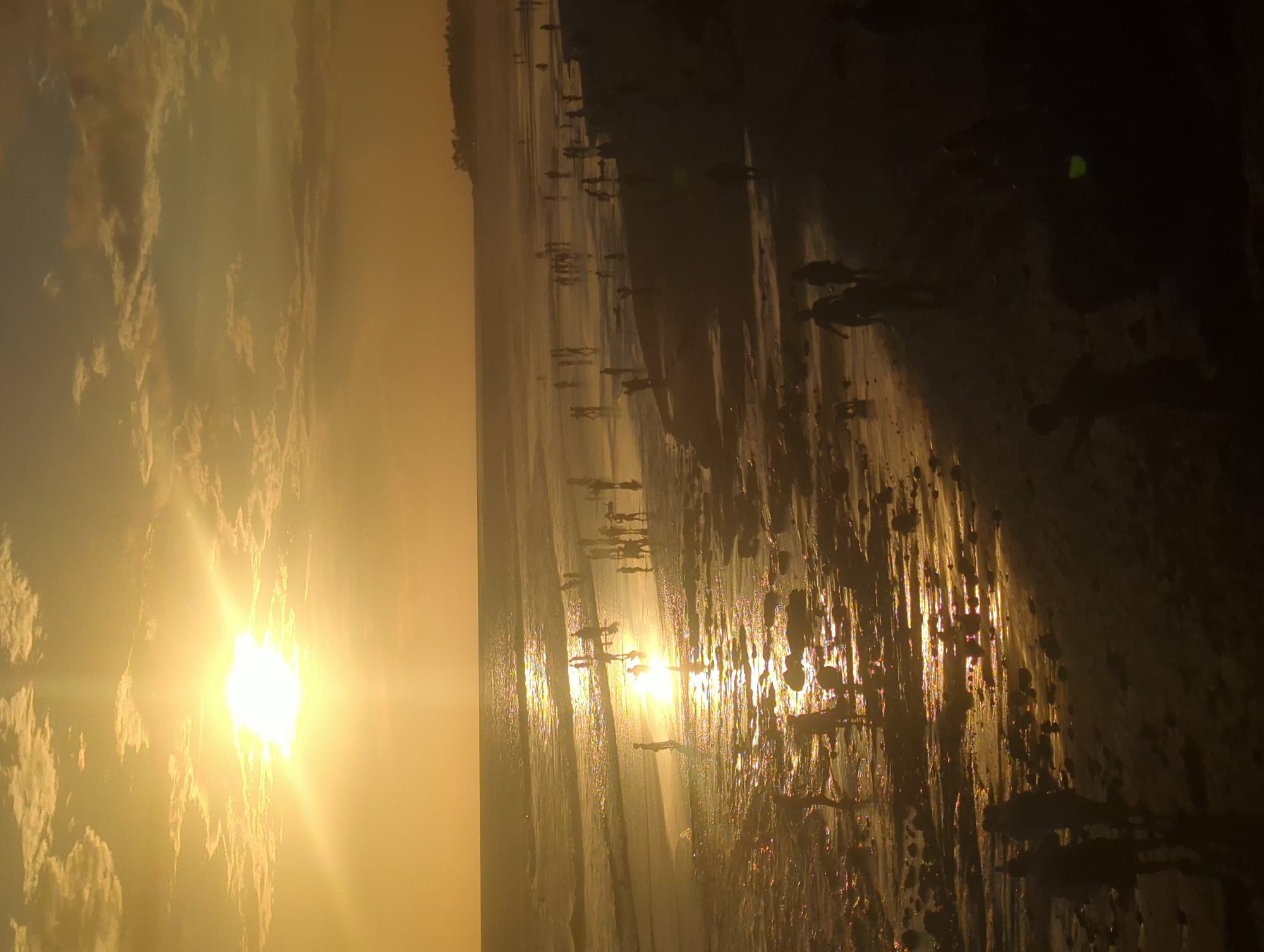 The image depicts a sunset at a beach in North Kuta, Indonesia. The sun is a bright orb in the sky, partially obscured by clouds. Its reflection is visible in the wet sand and shallow water along the shoreline. Numerous people are present on the beach, their silhouettes appearing darker against the bright sky and water. The environment consists of a sandy beach, the ocean, and the sky. The people appear to be wading in the water or standing along the shoreline.