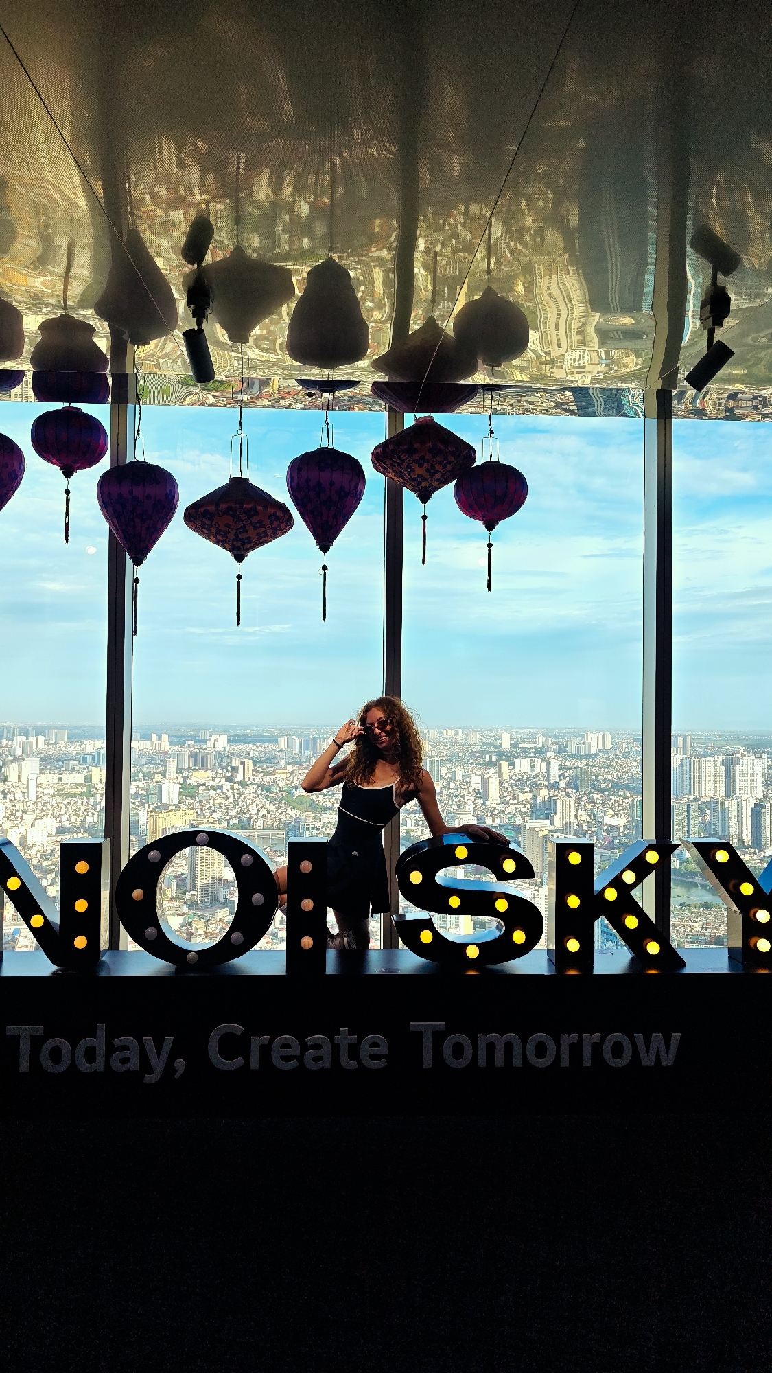 A woman with curly, blonde hair and sunglasses poses for a photo in front of a panoramic city view. She is wearing a black tank top and skirt, with one hand on her hip and the other resting on a large, illuminated letter sign that spells "NOISKY". The sign is composed of individual, marquee-style letters with yellow lights. Above the sign, several ornate, Asian-style lanterns hang from the ceiling, which reflects the city lights and the lanterns themselves. The woman appears to be cheerful and relaxed.
The backdrop is a cityscape with numerous buildings stretching to the horizon, suggesting a large, urban environment. The sky is a clear blue with scattered clouds, indicating daytime. The overall scene suggests the photo was taken at a high vantage point, likely a skyscraper observation deck or a rooftop bar.
Below the "NOISKY" sign, a second, darker sign reads "Today, Create Tomorrow" in grey, block lettering. The presence of the "NOISKY" sign and the cityscape of Hanoi strongly suggests the location is in Hanoi, Vietnam. The time of day appears to be late afternoon or early evening, as the sky is bright but the city lights are beginning to twinkle.