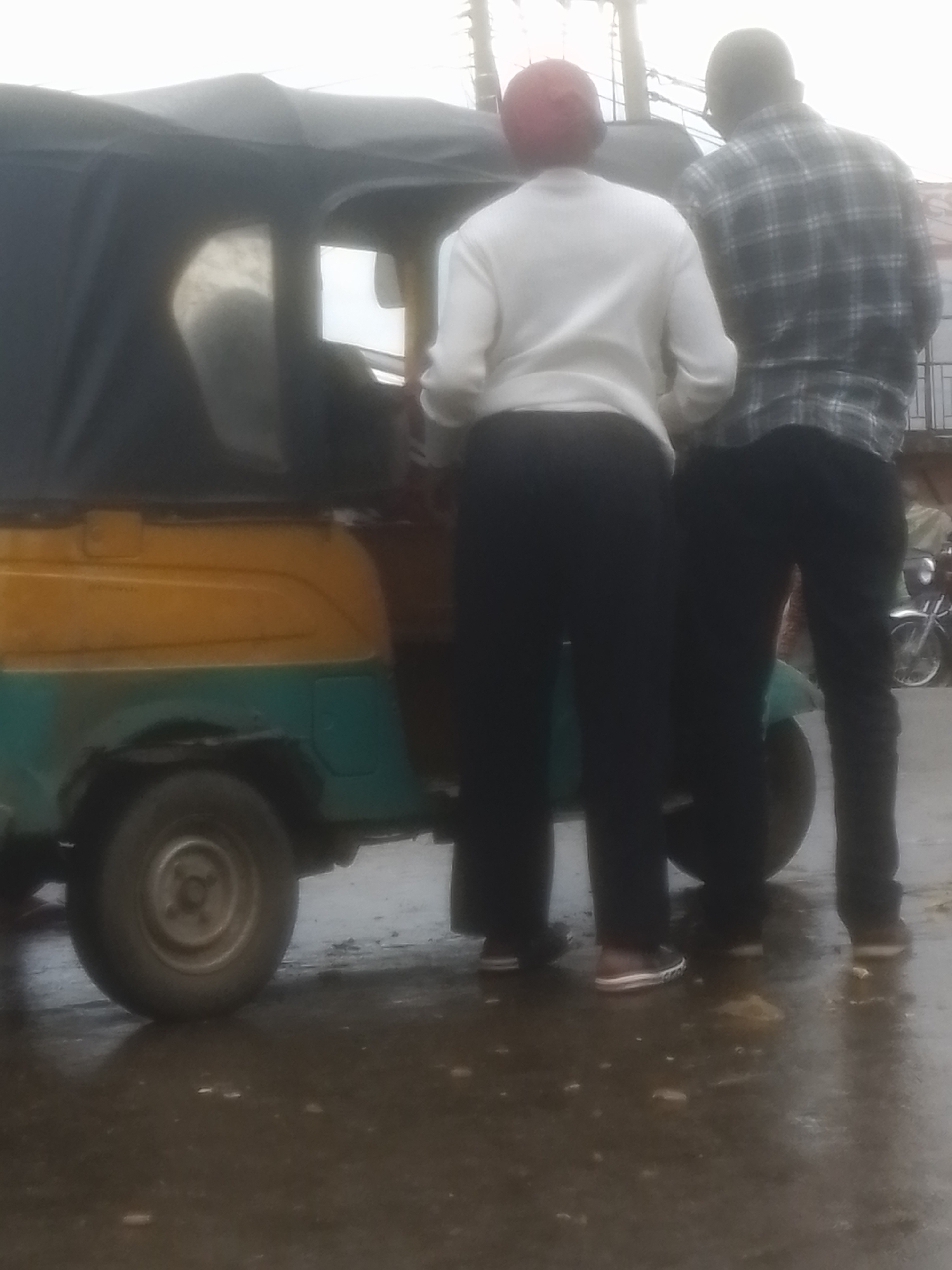 Two individuals are standing next to a three-wheeled vehicle, possibly an auto-rickshaw, in Gwong, Nigeria. The individual on the left is wearing a white long-sleeved shirt and black pants, with a red cap. The individual on the right is wearing a plaid shirt and black pants. The auto-rickshaw is yellow and teal. The ground is wet, indicating it may have rained or is raining. The scene appears to be outdoors, possibly on a street or in a marketplace.
