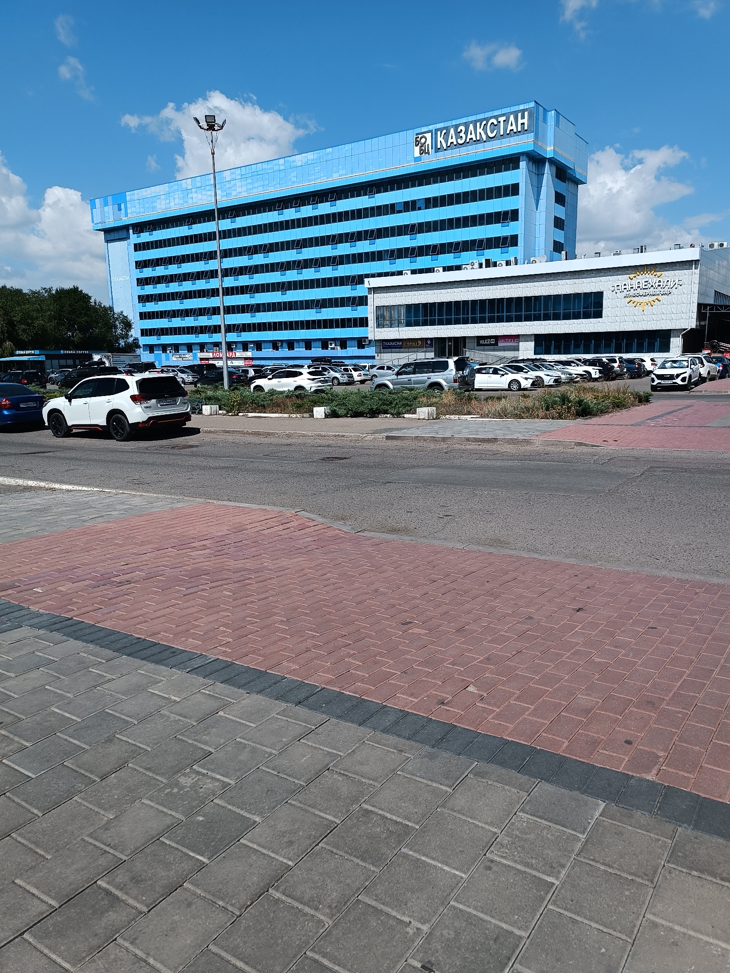 The image captures an exterior view of a modern building complex under a bright, partly cloudy blue sky. The main structure is a tall, multi-story building with a prominent blue facade and numerous windows. Above the windows, "ҚАЗАҚСТАН" (Kazakhstan) is displayed in large, white lettering against a blue background. Adjacent to this main building is a lower, white structure with a gray, textured facade, featuring large windows and signage that includes a sunburst emblem and text.

In the foreground, a paved area is visible, with a section of reddish-brown brick pavers transitioning to gray concrete pavers. A road runs horizontally across the middle ground of the image, with several vehicles parked or driving on it. A white SUV is particularly noticeable in the left-center of the frame, with its rear facing the camera. Other cars, including sedans and SUVs, are parked in a lot to the right of the road, in front of the white building. Green bushes and trees are present along the edge of the parking lot and behind the building on the left. A tall lamppost with multiple lights stands near the main building. The overall impression is of a well-maintained, commercial or administrative area in a city.