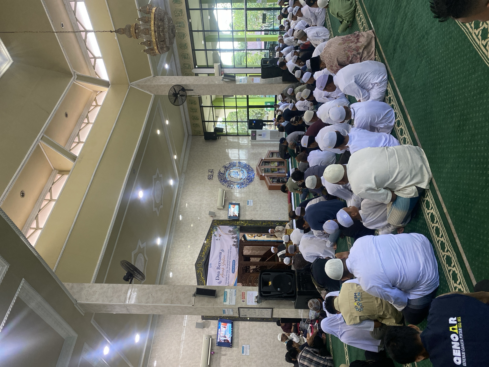 The image shows an indoor gathering of people, likely at a mosque, based on the attire of the individuals and the presence of religious decorations. A banner at the front reads "Doa Bersama Anak Yatim" (Prayer Together with Orphans) and specifies the event date as "Selasa, 29 Juli 2019" (Tuesday, July 29, 2019). The individuals are predominantly male, many wearing peci hats or sarongs. They are seated on a green carpeted floor. In the foreground, a person wearing a dark blue shirt with white text "QENOAR GENERASI CAHAYA PINTAR KAL-SEL" is visible. Behind them, many people are seated in rows, facing a raised platform or stage where several individuals are standing and speaking. There are screens displaying images on the walls, and a large chandelier hangs from the ceiling. The architecture includes columns and a high ceiling with exposed beams and lighting fixtures. The overall scene suggests a religious event or prayer service. The text "KAL-SEL" on the shirt in the foreground is likely an abbreviation for Kalimantan Selatan, which is South Kalimantan province in Indonesia, and Banjarbaru is a city within this province. Thus, the event is likely taking place in Banjarbaru, Indonesia.