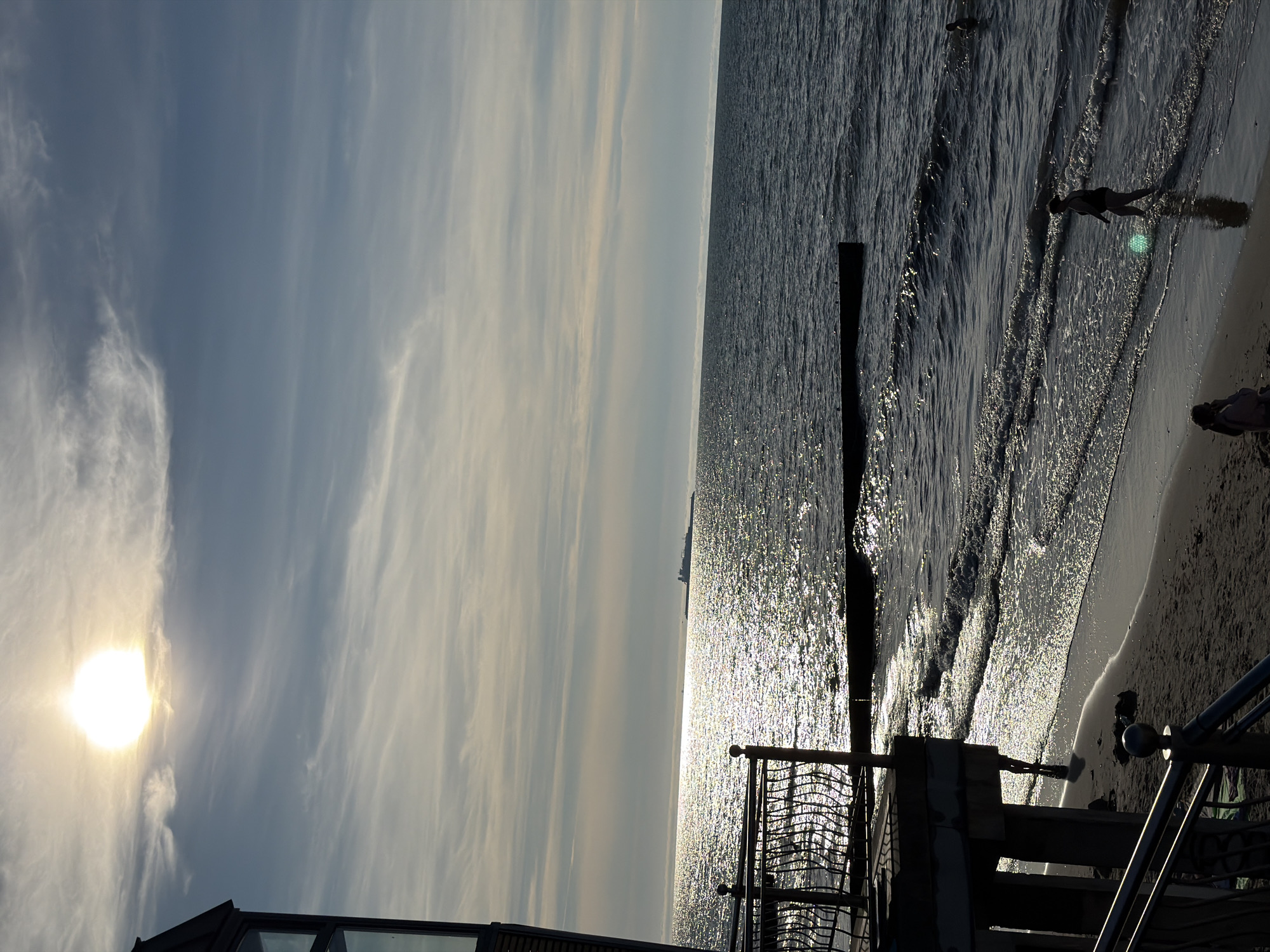 The image depicts a sunlit beach scene in Zelenogradsk, Russia. In the foreground, a dark, angular structure with windows extends from the left side of the frame, casting a shadow over the sandy beach and the edge of the sea. A decorative metal railing runs along the edge of this structure and continues down towards the beach.
On the beach, several individuals are visible. One person is walking into the shallow water, their silhouette dark against the bright reflection of the sun. Further out in the sea, another person can be seen partially submerged in the water. The waves gently lap the shore, creating a shimmering effect on the sand and in the water due to the sunlight.
In the distance, across the expansive sea, a large ship is visible on the horizon. The sky above is a pale blue, with wispy clouds streaked across it, some illuminated by the bright, diffused sunlight. The overall impression is one of a calm, late afternoon or early evening by the sea.
