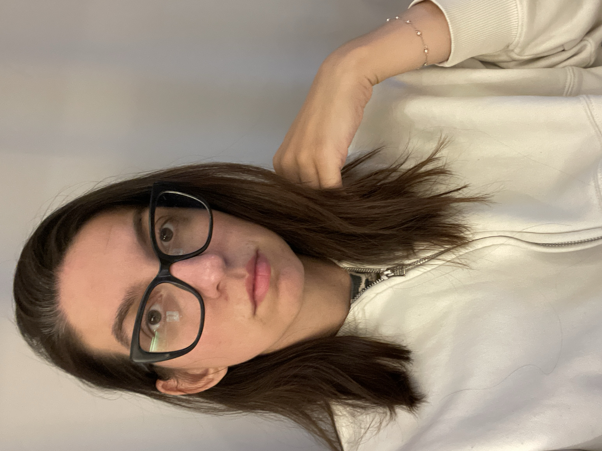 A young woman with brown hair and glasses is posing for a close-up selfie. She is wearing a light-colored zippered sweatshirt and a delicate bracelet is visible on her wrist. The background is a plain, light-colored wall. There is no information in the image to indicate the location is Sofia, Bulgaria.