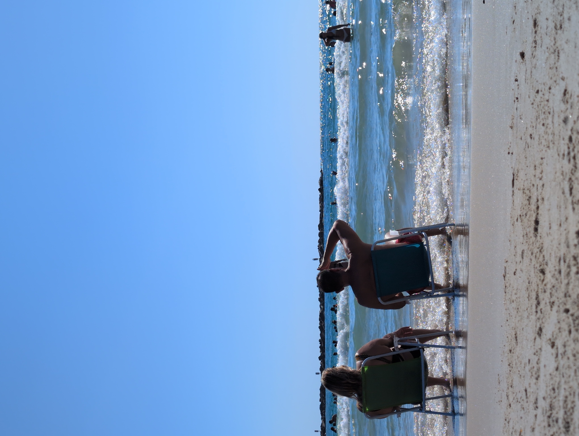 Two individuals are seated in beach chairs on a sandy beach, facing the ocean. The person on the left, viewed from the back, has blonde hair and is wearing a dark-colored swimsuit with a patterned top. The person on the right, also viewed from the back, has short dark hair and is wearing red swimming shorts. They are both looking out at the water. The ocean is blue with visible waves and sunlight glinting off the surface. Several other people are in the water, swimming and wading. In the distance, a breakwater structure made of large rocks extends into the sea, with navigational buoys visible. The sky above is clear and blue. The overall scene depicts a bright, sunny day at a beach. This scene is likely at one of Tel Aviv's popular beaches such as Gordon Beach or Frishman Beach, given the presence of beach chairs, breakwaters, and numerous bathers.