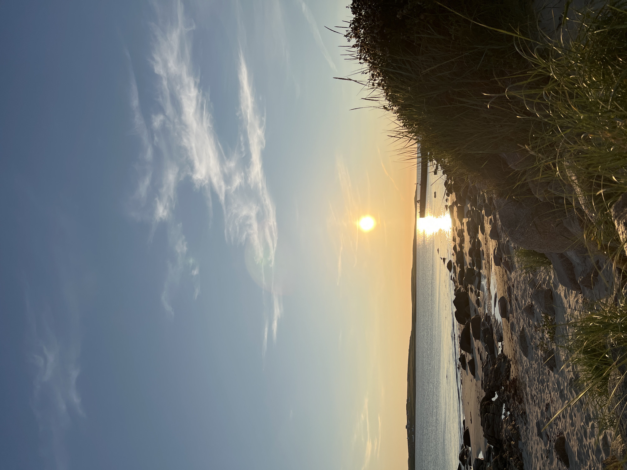 The image depicts a serene coastal scene at sunset in Lunniaghbeg, Ireland. The sun, a bright orb, hovers just above the horizon, casting a warm golden glow across the sky and reflecting a shimmering path on the calm sea. Wispy clouds streak across the pale blue sky, transitioning to hues of orange and yellow near the sun.
The foreground is a sandy beach littered with rocks of varying sizes, some smooth and rounded, others jagged and dark. Patches of coarse green grass and reeds grow amongst the rocks and in the dunes, with some blades of grass catching the late afternoon sun. In the distance, a low, green island with a few buildings is visible across the water, appearing as a dark silhouette against the bright sky. A long pier or breakwater extends from the island into the sea. The overall impression is one of tranquility and natural beauty.