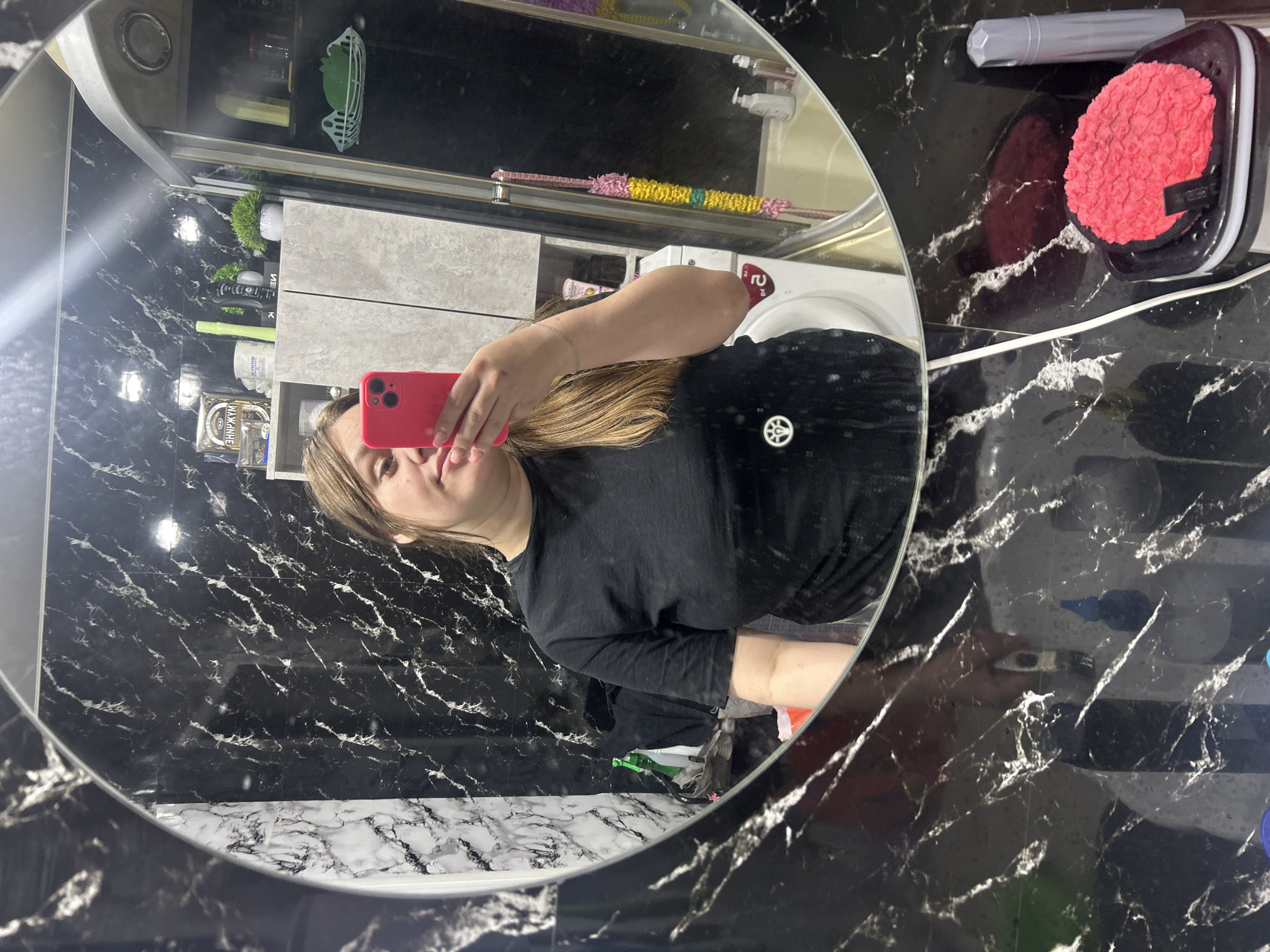 A female individual is depicted taking a selfie in a bathroom mirror in the city of Ольховка, Russia. The individual, seen from the chest up, has light brown hair and wears a black short-sleeved t-shirt with a small white emblem on the left side. She holds a red smartphone with a triple camera array in her raised left hand, with a silver bracelet visible on her wrist. The bathroom features black walls with prominent white marble-like veining. Reflected in the mirror behind the individual are: a light grey two-door cabinet holding various personal care items and small potted plants, a partially visible white washing machine, and a shower enclosure or bathtub. Inside the shower, a light blue wall-mounted basket holds a green item, and various pump bottles are visible on a shelf or ledge. The vanity surface in the immediate foreground, outside the mirror's reflection, also exhibits black marble patterning and contains a white power cord, a black tray holding a bright red textured circular object, and a grey cylindrical container.