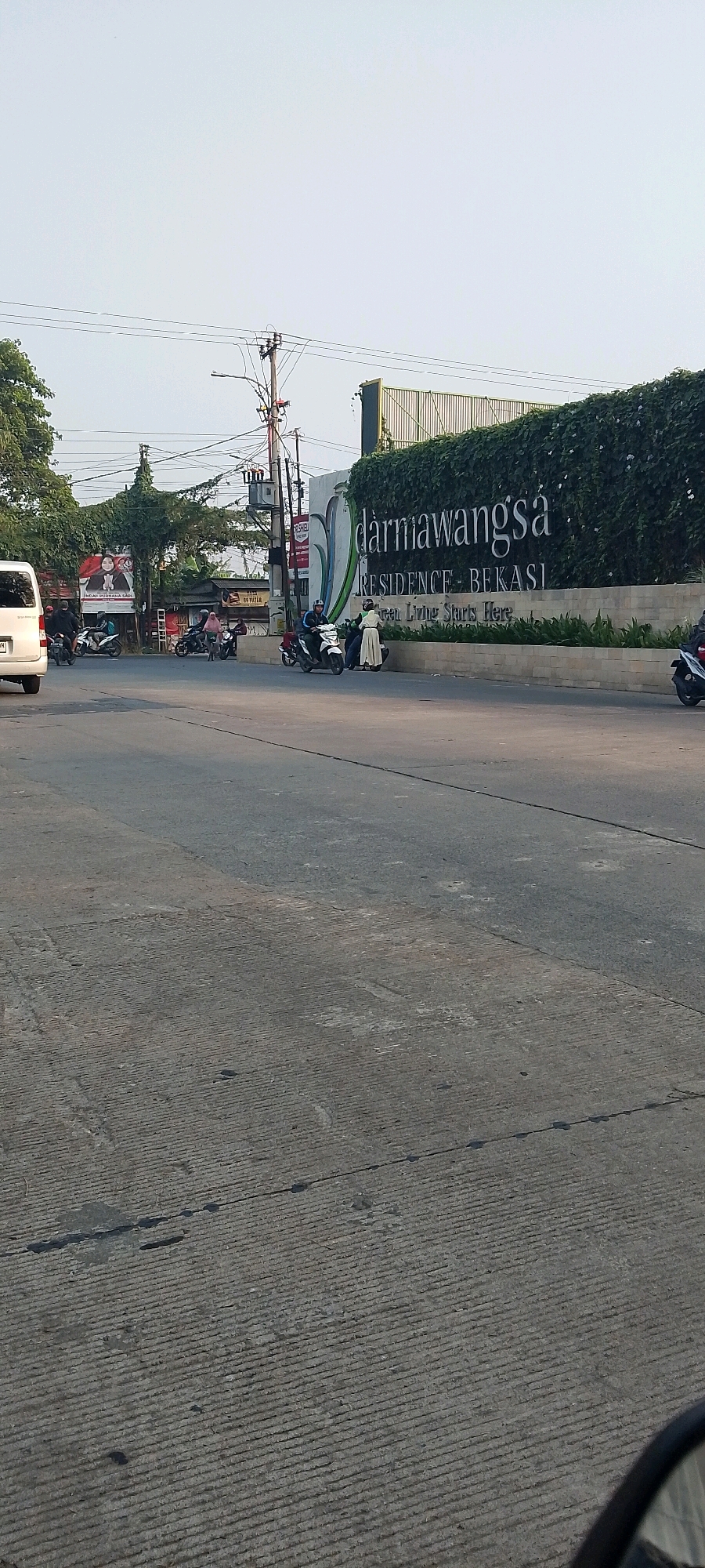 A street scene is captured in Satriamekar, Indonesia, featuring a prominent sign for "darmawangsa RESIDENCE - BEKASI" with the tagline "Green Living Starts Here." The sign is adorned with lush green foliage. In the foreground, a paved road with distinct grooved texture is visible, indicating wear and tear. To the left, a white van is parked. Motorbikes are seen on the road, with riders engaged in travel. Several individuals are present, some on motorbikes and others standing. The environment suggests an urban setting with overhead power lines and utility poles. The sky is clear and light.