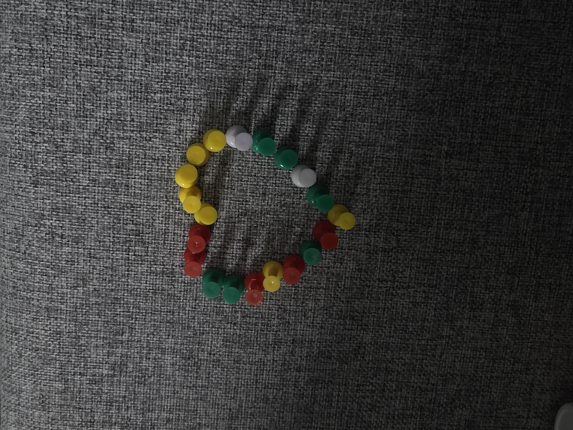 On a textured gray surface, an incomplete, irregular heart shape is formed by an assortment of round pushpins. The pushpins are colored yellow, white, green, and red. The location is unknown.