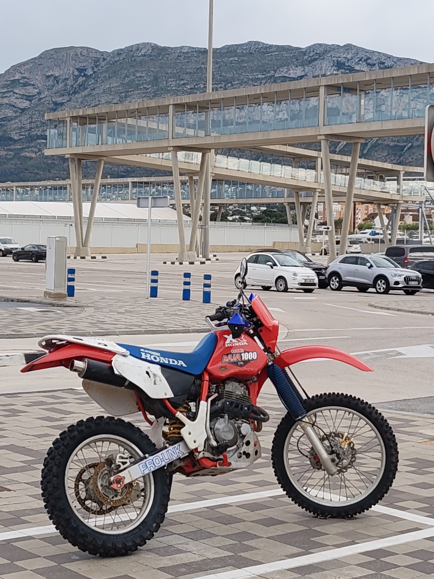 The image shows a red, white, and blue Honda Baja 1000 motorcycle parked on a checkered pavement surface in what appears to be a parking area. Several cars, including a white Fiat 500 and a silver Audi Q3, are visible in the background. Behind the cars is a modern, elevated walkway or building structure supported by multiple columns. In the distant background, there are mountains. The location is Dénia, Spain.