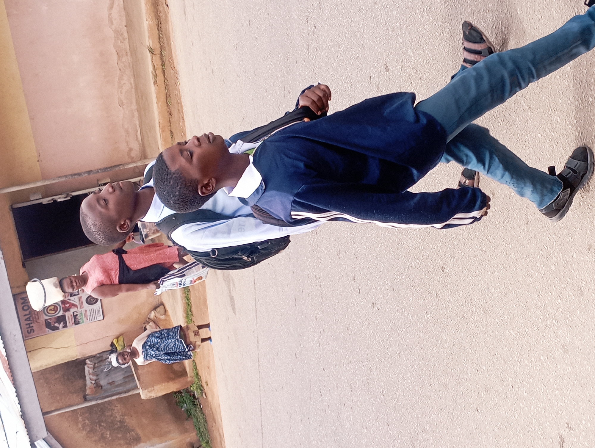The image depicts a street scene in Jos, Nigeria, focusing on three individuals walking on a paved road. In the foreground, two young males are walking from left to right. The one closer to the camera is wearing a navy blue jacket with white stripes on the sleeves, a white collared shirt, and blue jeans. He is also wearing dark sandals. Behind him, another young male is visible, also wearing a white collared shirt and a backpack. In the background, two women are standing near a building. One woman is wearing a pink dress and carrying a bucket on her head and a bag in her hand, while the other woman, standing to her left, is wearing a blue and white patterned garment and a headscarf. The building behind them has signage that reads "SHALOM." The environment appears to be an urban or semi-urban setting with a muted color palette, suggesting an overcast or dusty day.