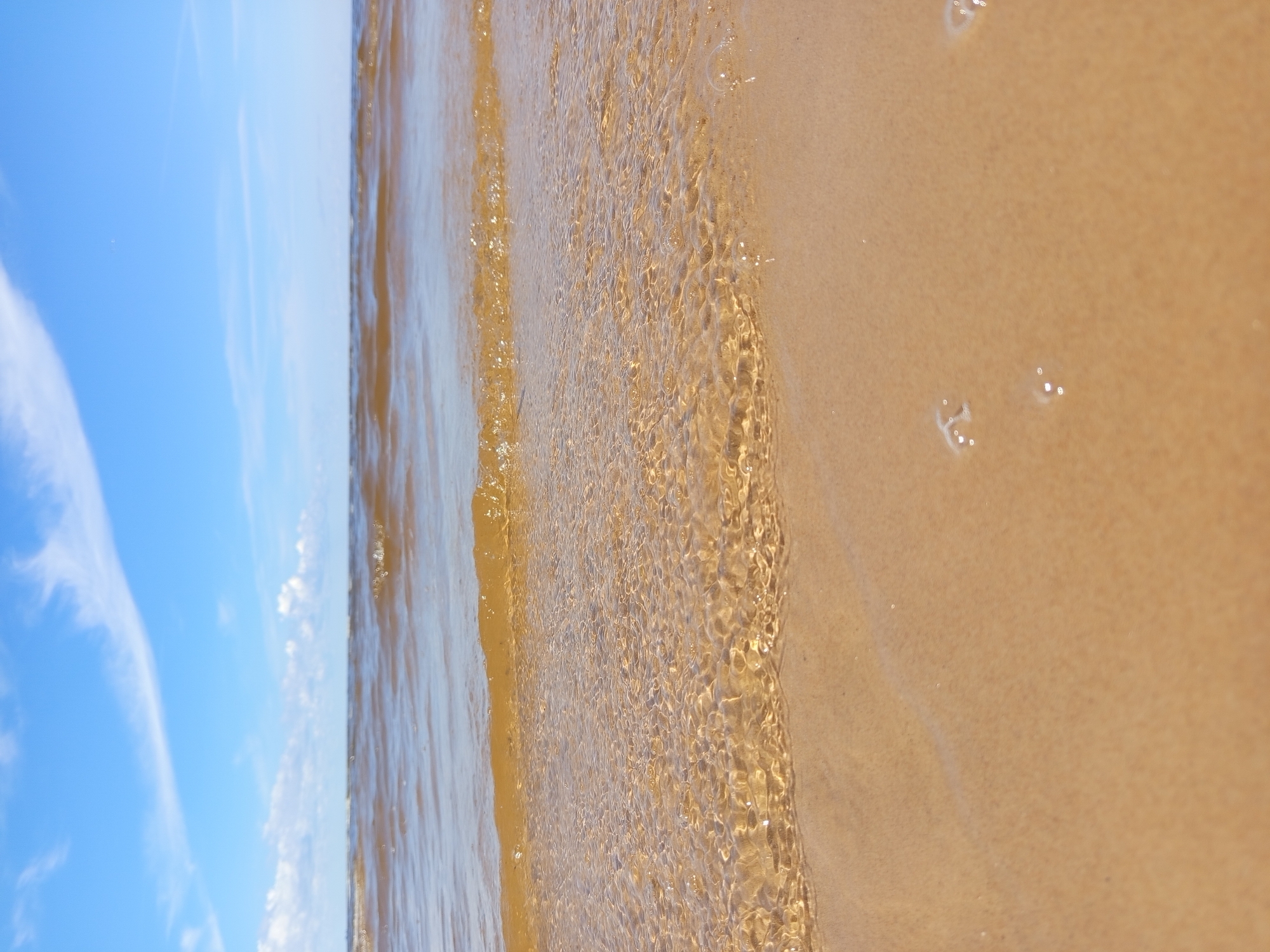 The image shows a sandy beach with calm, shallow water lapping at the shore under a bright blue sky with scattered white clouds. The water is a murky brown color, and small waves are creating ripples on the sandy bottom. There are a few small bubbles on the wet sand in the foreground. The horizon line is visible in the distance, separating the water from the sky. The scene is natural and appears to be a peaceful coastal environment. There are no people or man-made objects visible in the frame.