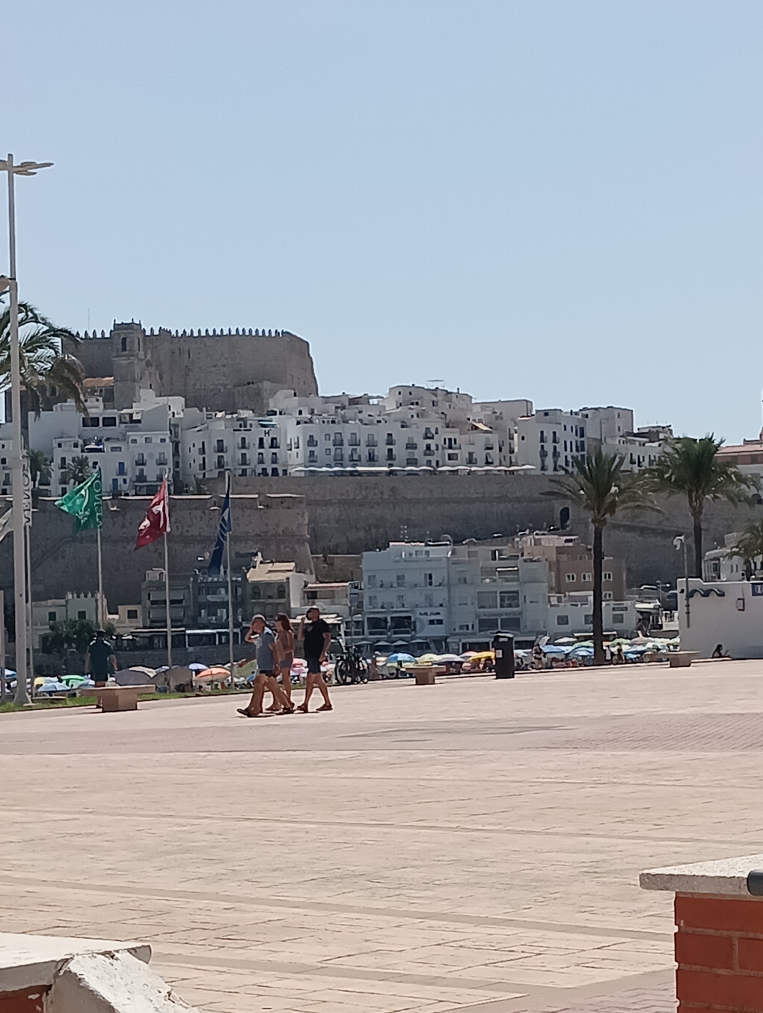 The image shows a view of the city of Peníscola, Spain, dominated by the Castillo de Peñíscola, a large fortress situated on a rocky promontory. The white buildings of the city cascade down the hillside towards the sea, with numerous windows and balconies visible. In the foreground, a paved plaza area is occupied by a few people walking. Three flags on flagpoles are visible, one green, one red with a crest, and one blue. Palm trees are scattered throughout the scene. In the middle ground, beach umbrellas and lounge chairs are set up, indicating a beach or promenade area. The sky is clear and blue, suggesting a sunny day.