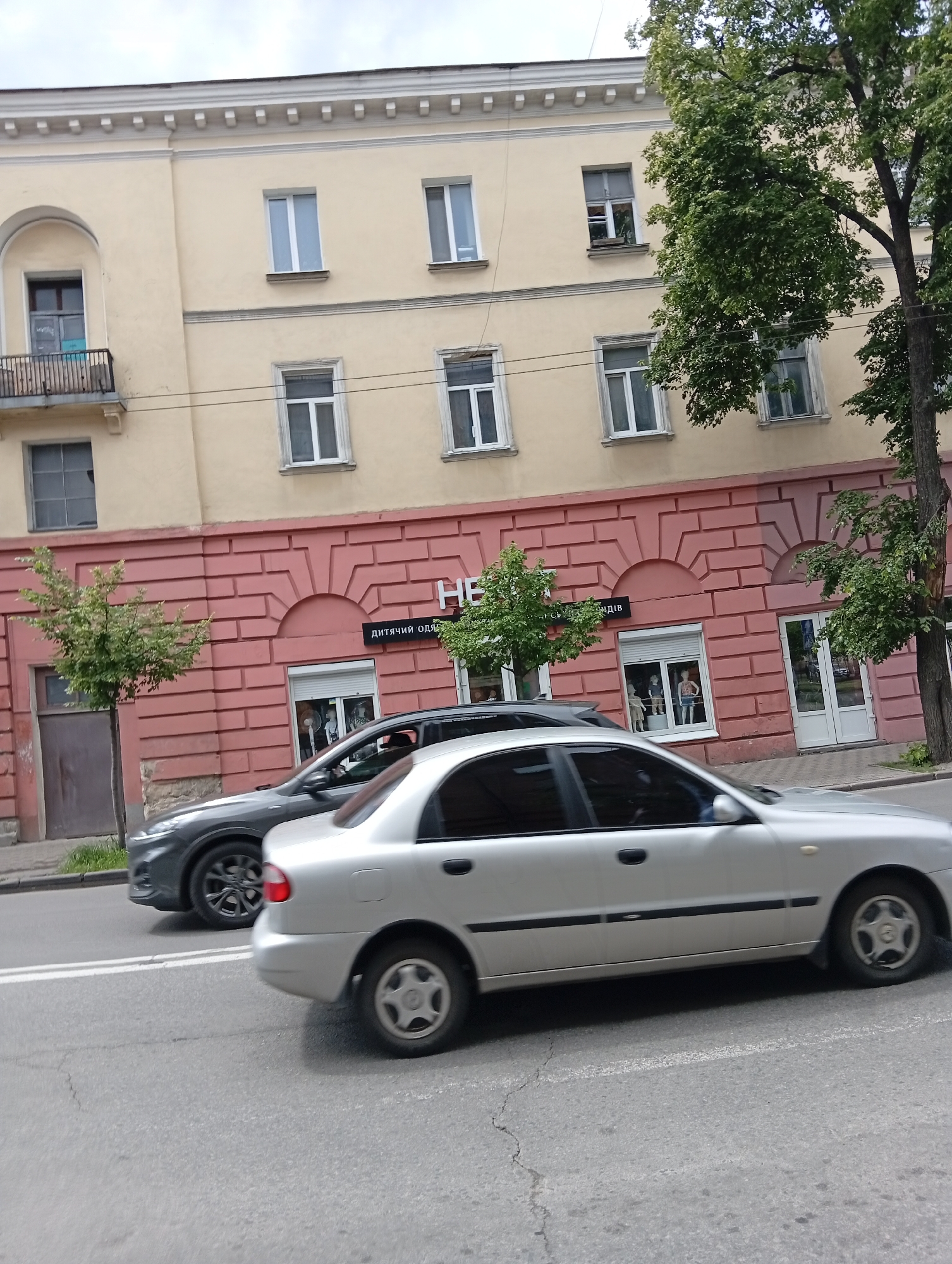 The image shows a street scene in Poltava, Ukraine. A silver Daewoo Lanos is driving on the road, passing a gray Hyundai Kona. In the background is a multi-story building. The ground floor is red with a brick pattern and houses a shop named "Дитячий Одяг" (Children's Clothing). The upper floors are painted yellow and have white-framed windows. Trees line the street. The sky is overcast.