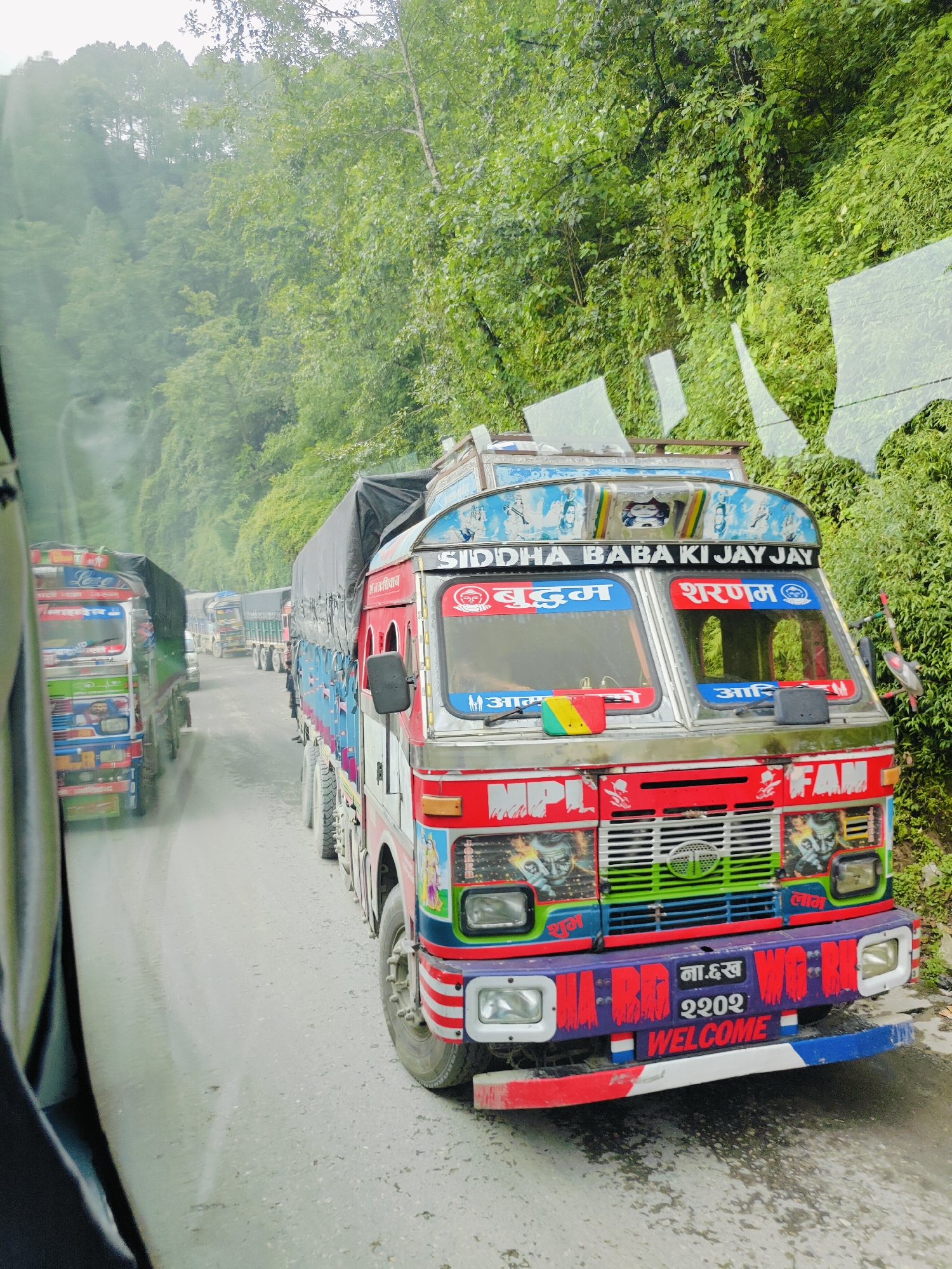 The image depicts a traffic jam on a wet, paved road, likely in Chandragiri, Nepal, bordered by lush green foliage and trees on a steep incline. Several brightly decorated trucks are lined up, their colorful paintwork contrasting with the overcast sky. The front truck, a red Tata model, is heavily adorned with religious imagery, text in Hindi and English, and graphics. Visible text includes "SIDDHA BABA KI JAY JAY," "बुद्धम," "शरणम," and "WELCOME." Other trucks in the convoy also exhibit vibrant decorations. The wet road surface and the presence of mist or light rain suggest recent or ongoing precipitation. The perspective suggests the photo was taken from within another vehicle.