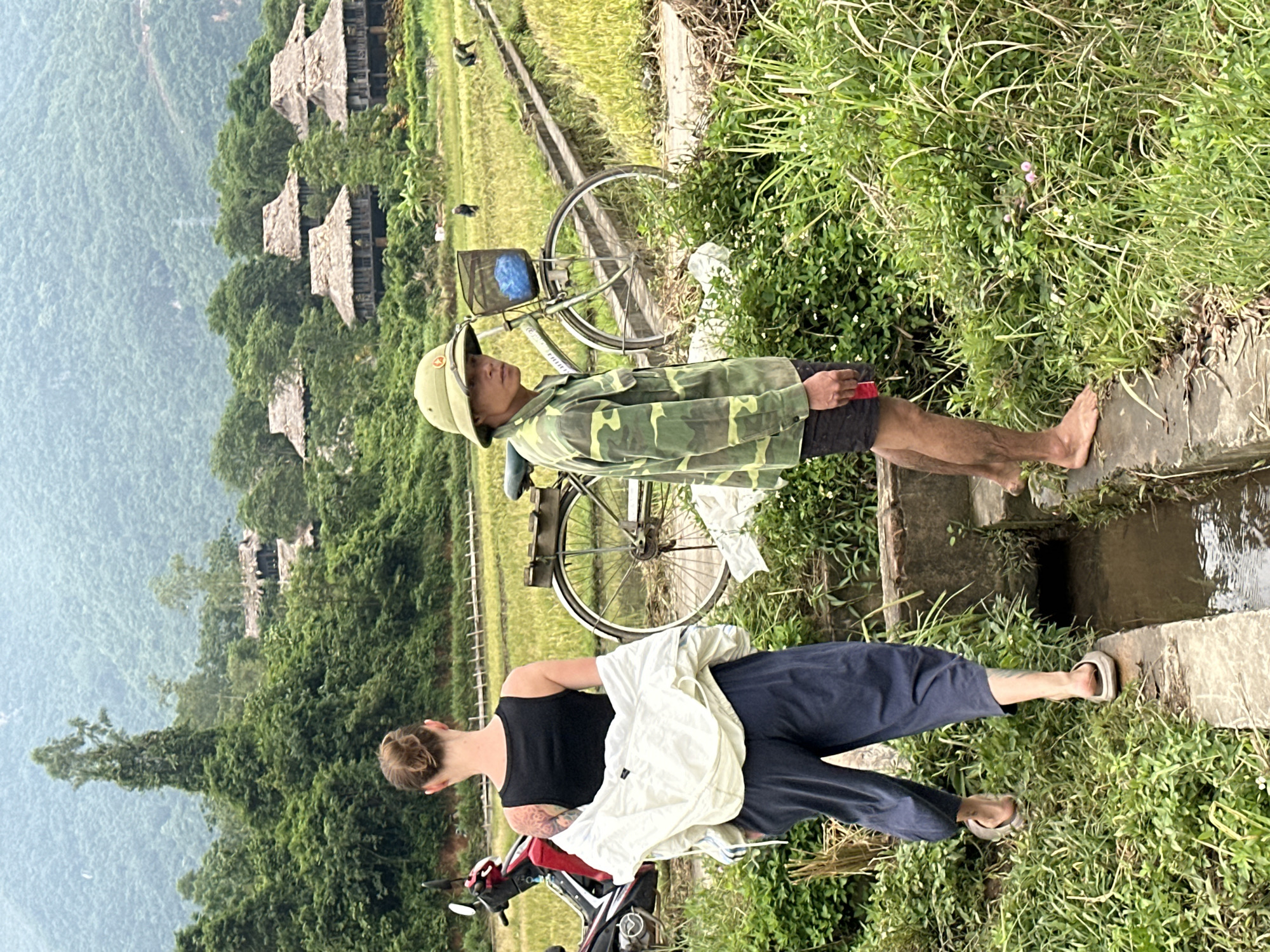 In Mai Chau, Vietnam, a man wearing a camouflage shirt and hat stands near a rice paddy. He is barefoot and holding a red object. Next to him, a woman with tattoos on her arm walks away from the camera, carrying a white jacket. A bicycle is parked behind them, with a blue basket on the handlebars. In the background, traditional stilt houses with thatched roofs are nestled among lush green trees, with rolling hills in the distance. A motorcycle is parked to the left of the woman.