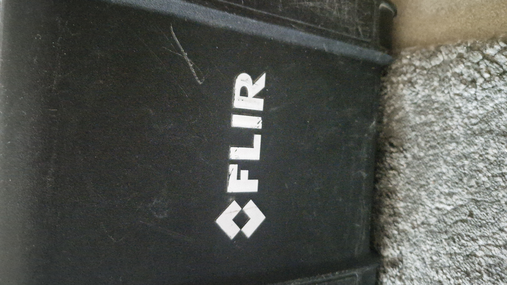 The image shows a close-up of a black object, likely a case or enclosure, with a white logo that reads "FLIR." The logo is centrally located and appears to be slightly worn. The object has a textured surface, and there are visible scratches and scuff marks on its surface, suggesting it has been used. The object is resting on a grey, carpet-like material. There is no discernible information about the location in Baden-Baden, Germany.