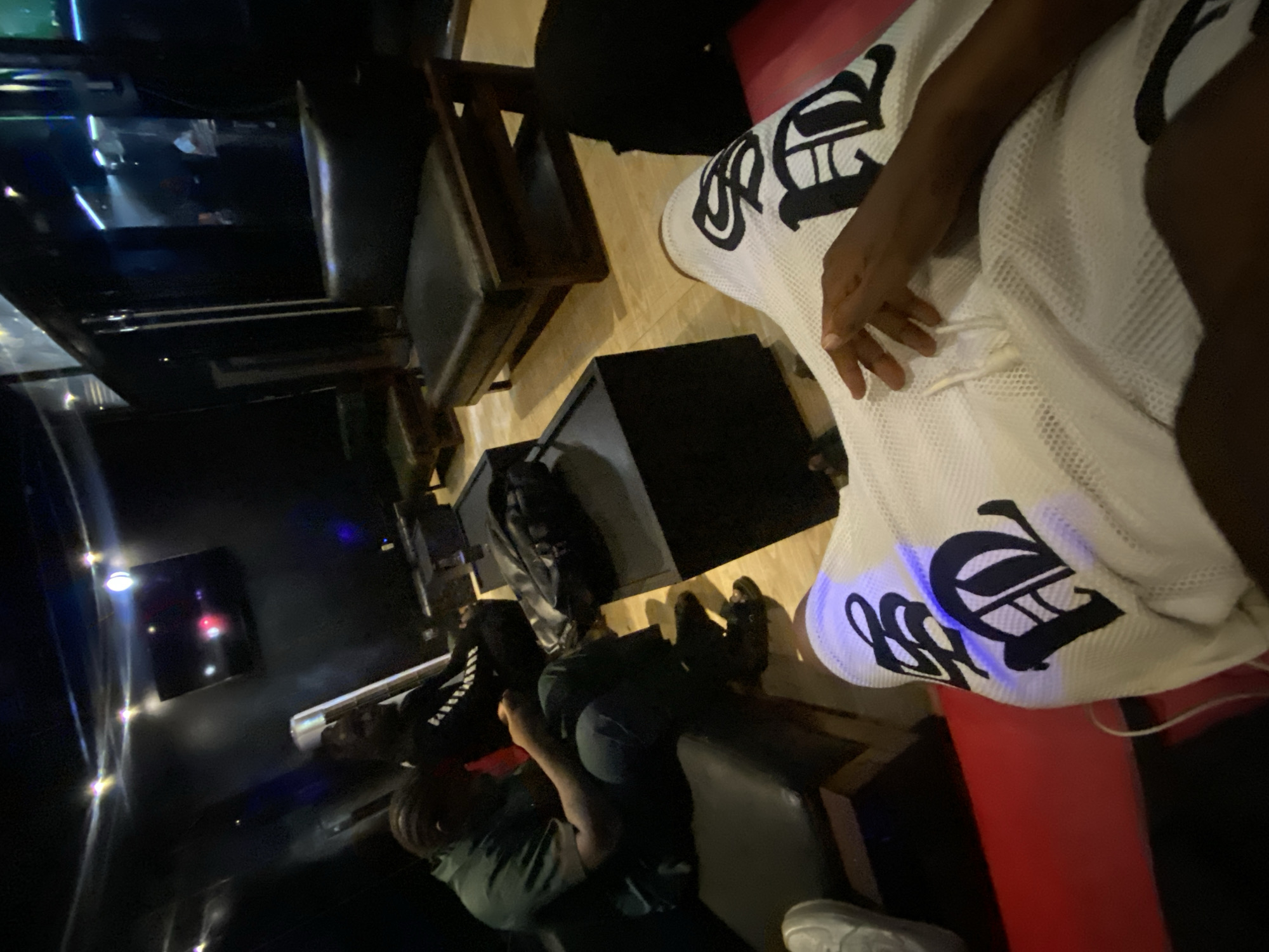 The image depicts an indoor setting in Iranla, Nigeria, possibly a lounge or entertainment venue. Two people are visible, one wearing a green shirt and another wearing a black shirt with white letters, both seated in upholstered chairs. A person wearing white shorts adorned with large black lettering sits on a red cushioned bench. In the background, there is a wall-mounted television and ambient lighting. Additional furniture includes wooden chairs with dark upholstery.