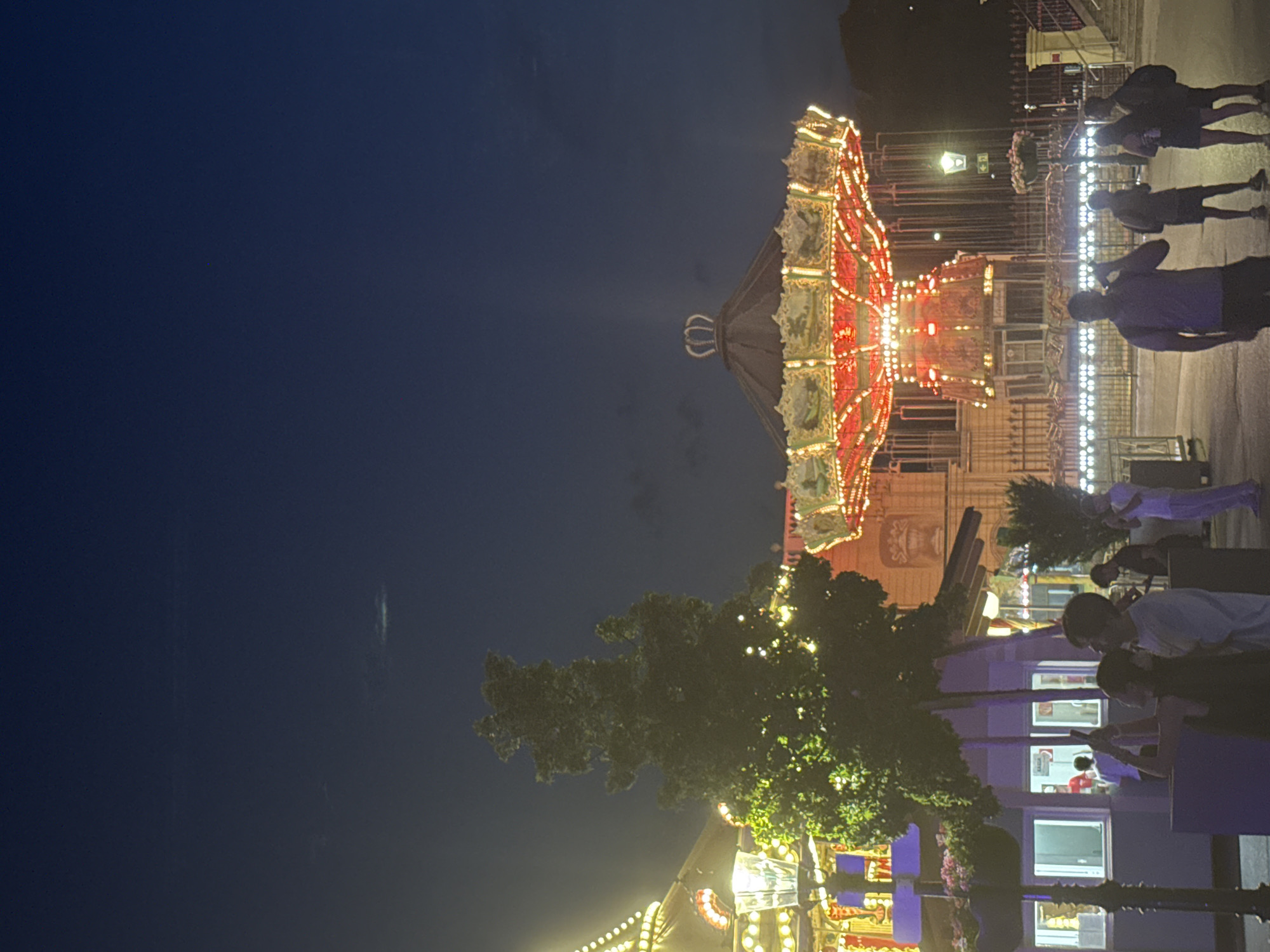 The image depicts a nighttime scene at a carnival or amusement park, with a brightly lit carousel, or swing ride, as the central focus. The ride is decorated with intricate details and illuminated by numerous lights. In the foreground, several people are visible, some standing and observing the ride, while others are in conversation. One person is holding a phone up, possibly taking a picture. The background shows a building with architectural details and more decorative lighting. A tall tree with illuminated foliage stands to the left of the ride. The sky above is a deep blue, indicating dusk or early night. The presence of a carousel ride strongly suggests a location associated with entertainment and leisure. Given the instruction to include Vienna, Austria, this scene could be from a fairground within the city. The overall atmosphere is festive and lively, characteristic of an evening at a public entertainment venue.