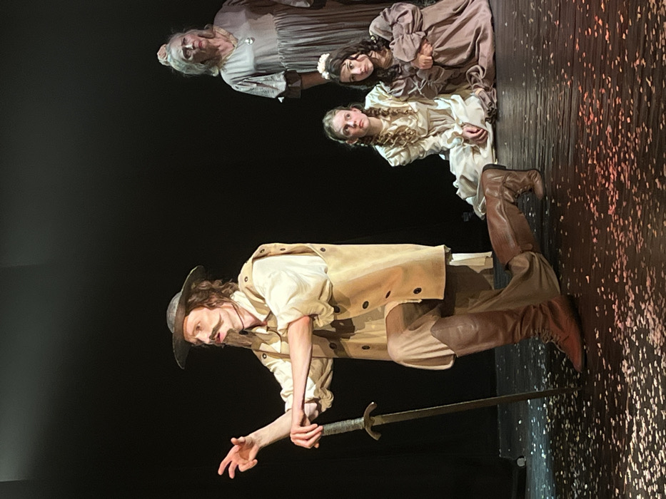 The scene depicts a theatrical performance taking place on a stage with a stark black backdrop in Novoshakhtinsk, Russia. In the foreground, a male actor, characterized by a long mustache and goatee, a wide-brimmed hat, light-colored shirt, tan tunic, and dark knee-high boots, kneels on his right knee. His left hand rests on the pommel of an upright sword, its tip on the confetti-strewn dark stage floor, while his right arm is raised and extended in a dramatic gesture. His gaze is directed off-frame to the left. To his right, two young females, dressed in period-style long dresses (one off-white, one light brown), sit on the stage, observing the male figure with serious expressions. Behind them, an elderly woman with light grey hair, wearing a loose grey dress and a necklace, stands partially visible, also looking towards the central male figure. The stage floor is dark, scattered with numerous small, light-colored paper fragments or confetti, illuminated by focused stage lighting.
