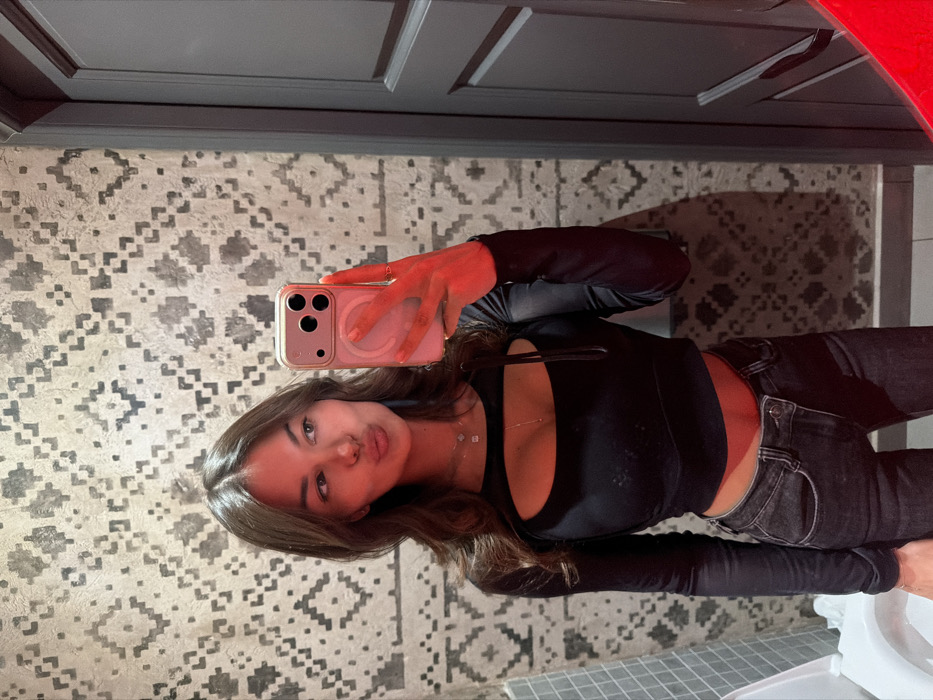 A young adult female is depicted taking a mirror selfie within a restroom in the city of Krasnoyarsk, Russia. She possesses long, dark brown hair, parted centrally, and presents with pursed lips while looking towards her smartphone. Her attire consists of a black long-sleeved crop top featuring a horizontal chest cut-out, paired with dark-wash jeans. Multiple delicate necklaces and a ring on her right index finger are visible. She holds a light-cased smartphone, consistent with an iPhone 14 Pro/Pro Max model with a visible MagSafe ring, in her right hand, angled towards the reflective surface. The background wall exhibits a textured surface with a repeating grey geometric pattern. To the right, a dark-colored paneled door is partially visible. On the lower left, a portion of a white ceramic fixture, likely a toilet or sink, is present. A distinct red light source illuminates the subject's right arm and the smartphone.
