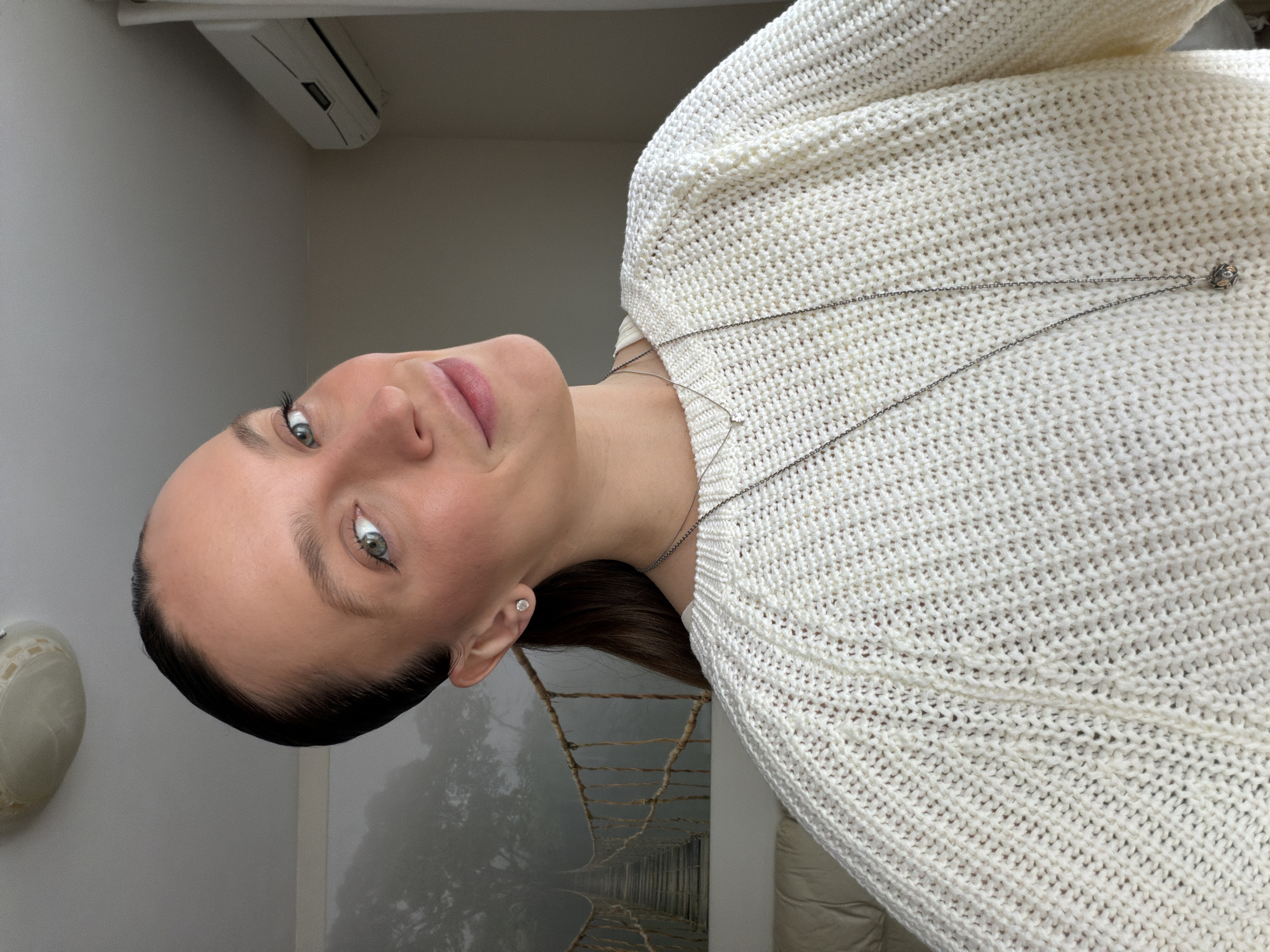 A light-skinned woman with dark hair and light eyes looks at the camera in an indoor setting. She wears a white knitted sweater and a silver necklace. Behind her is a wallpaper featuring a rope bridge surrounded by fog. The setting appears to be a room with neutral-colored walls. The location in Budva, Montenegro, cannot be determined from this image.