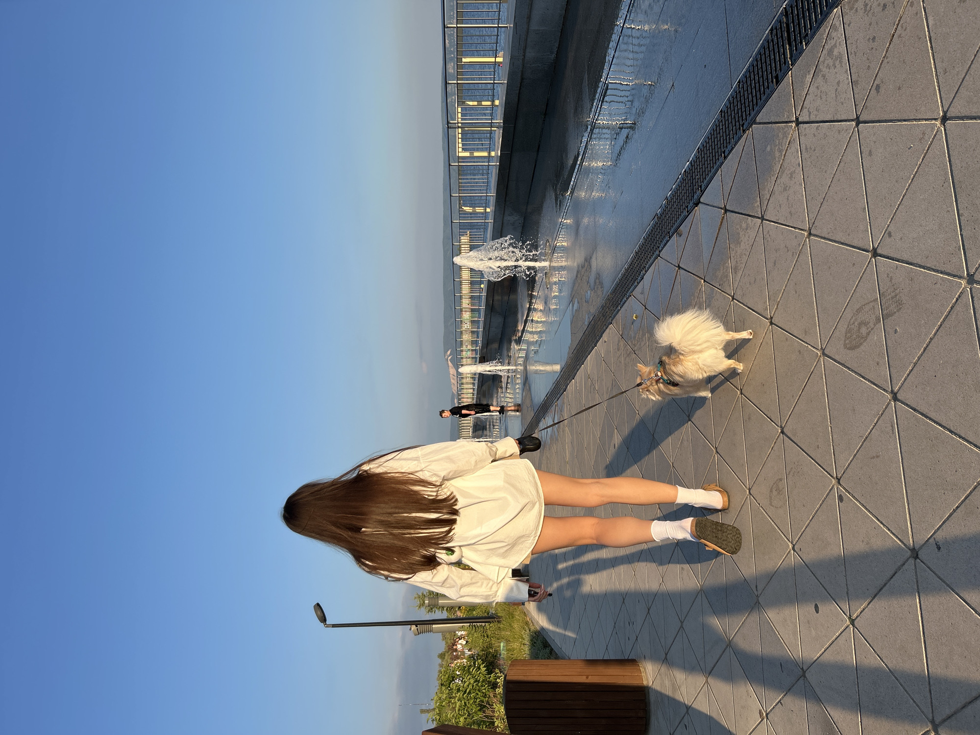 This image captures a serene outdoor scene in Tolyatti, Russia, likely during a warm, sunny day. The perspective is from behind a young woman with long, dark hair, wearing a loose white shirt and white socks with sandals. She is walking on a patterned tiled pathway next to a water feature with fountains. To her right, a small, fluffy light-colored dog on a leash walks alongside her. In the background, a metal railing lines a slightly elevated walkway, and further beyond, a body of water is visible, reflecting the sunlight. Another person can be seen walking in the distance along the elevated walkway. The clear blue sky suggests good weather. The overall mood is peaceful and relaxed, hinting at a leisurely stroll on a pleasant day. There is no visible text in the image.