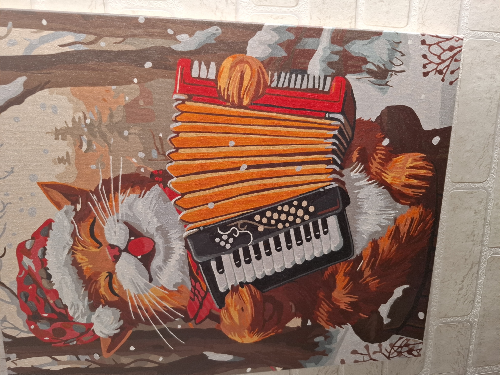 The image shows a painting of a sleeping cat with an accordion. The cat is ginger and white and is wearing a red scarf. The accordion is red, orange, and black. The background is a wintry scene with snow and bare trees. The painting is displayed on a wall with a light-colored brick pattern. The city of Амзя, Russia is not directly visible in the image.