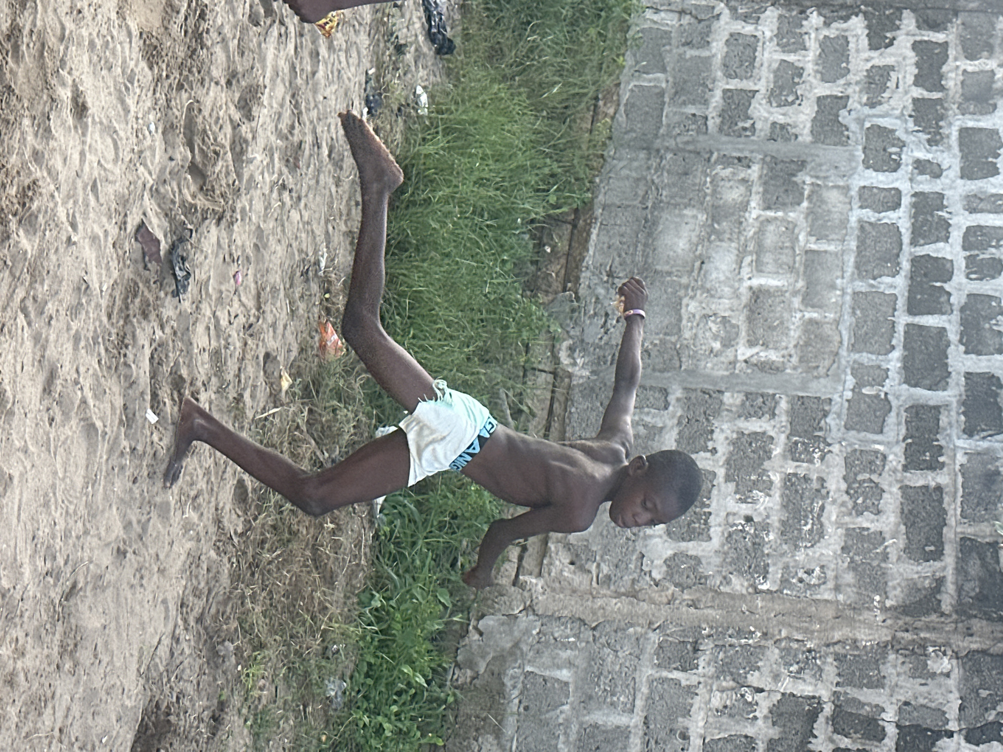 A young boy is performing a handstand on a sandy area next to a grassy patch and a concrete wall. He is wearing white shorts. The sandy area appears to have some scattered debris. The concrete wall is made of rough-hewn blocks. The scene is outdoors, and based on the lighting, it appears to be daytime. The boy's body is angled upwards, with his legs extended in the air. His face is turned downwards, and he seems focused on his balance. The overall impression is of a playful or athletic activity. The text "GRANGE" is visible on the waistband of the boy's shorts. The location is Badagry, Nigeria.