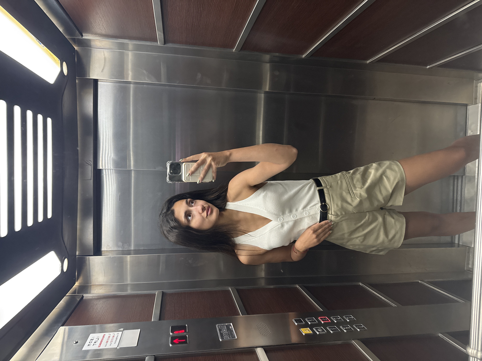 A young woman with long dark hair and fair skin stands in an elevator, taking a selfie. She is wearing a white sleeveless V-neck top with buttons down the front, and light beige high-waisted shorts with a black belt. The elevator's walls are a mix of stainless steel and dark wood panels. On the left, there is a panel with elevator buttons, including illuminated red up and down arrows, and a series of numbered buttons below. A sign with red text is affixed to the panel above the buttons. The lighting in the elevator is bright, emanating from long fluorescent fixtures on the ceiling. The overall environment suggests a modern building, possibly a hotel or residential complex in Budva, Montenegro.