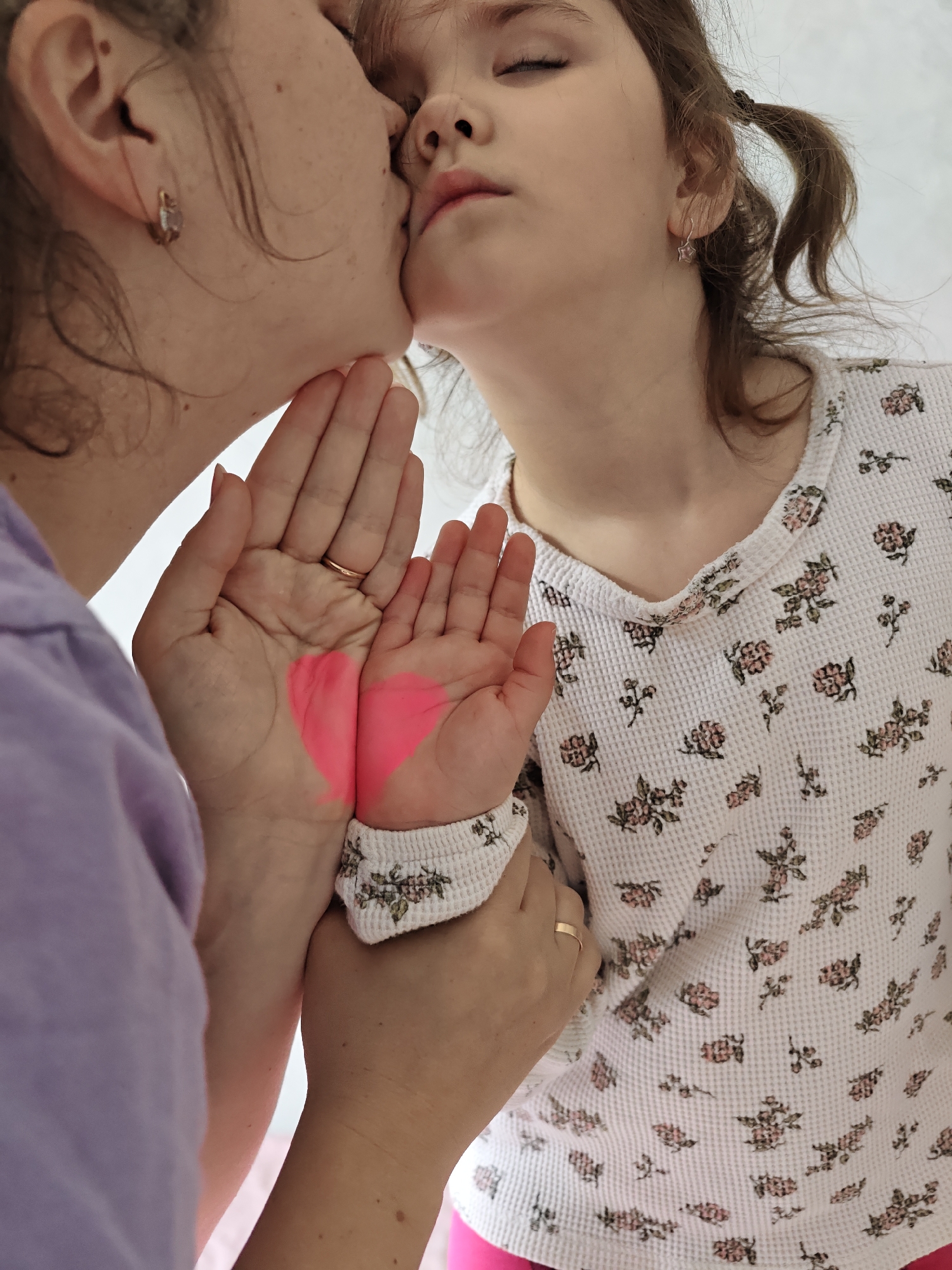 This intimate portrait captures an adult and a young girl, likely a mother and daughter, in a tender moment indoors in Chaikovsky, Russia. The adult, positioned on the left, gently kisses the child's cheek or temple with her eyes closed, conveying deep affection and warmth. She wears a light purple top, and a simple gold band ring is visible on her right ring finger. A small hoop earring can also be seen.
The child, on the right, receives the kiss with her eyes closed, her brown hair styled in pigtails. She is dressed in a white waffle-knit long-sleeved shirt patterned with small pink and green floral motifs, and appears to be wearing pink trousers or leggings. A small star-shaped earring is visible in her ear.
A central detail is the adult holding the child's hands, which are cupped together to prominently display a vibrant pink heart painted across both palms. This heart emphasizes the theme of love and connection between the two subjects. The background is plain and light, directing the viewer's focus entirely on the affectionate interaction. The overall scene radiates feelings of love, care, and intimacy. No visible text is present in the image.