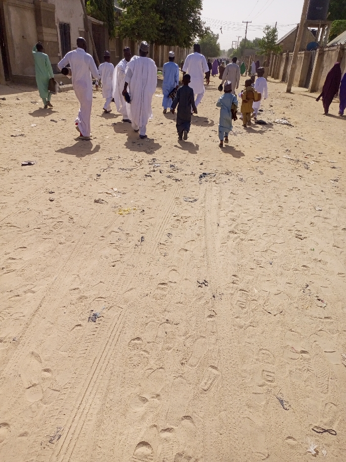 The image shows a group of people walking on a dirt road in Maiduguri, Nigeria. The road is covered in sand and has tire tracks. The people are all wearing traditional clothing and walking in the same direction. The environment is dry and dusty. The scene is captured from a high angle, looking down on the group as they walk away from the viewer. There is a building in the background to the left of the image. There are no trees to the right of the image, only a wall. The road is empty except for the people in the image, with some debris scattered on the ground.