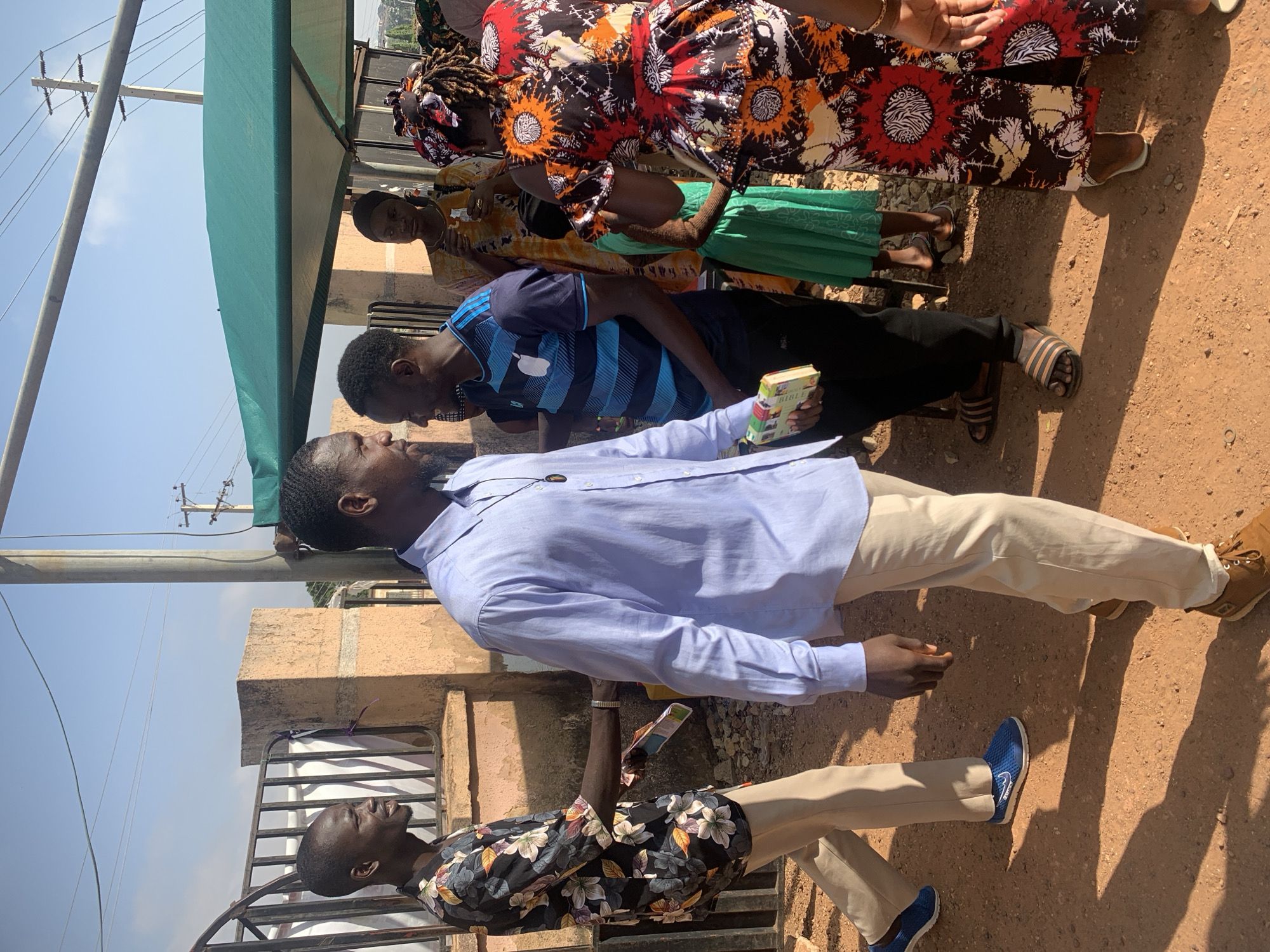 A group of people are standing outdoors in Jos, Nigeria, on a sunny day. The ground is reddish-brown. One man is holding a Bible and wearing a light blue long-sleeve shirt with khaki pants and brown shoes. He is facing another man in a blue striped shirt and dark pants, who is also holding a small book. Further behind them are women wearing colorful patterned clothing. One man is standing near a fence in a black floral shirt and tan pants. There's a green awning overhead, and a building is partially visible behind them. Electrical poles are visible in the background.