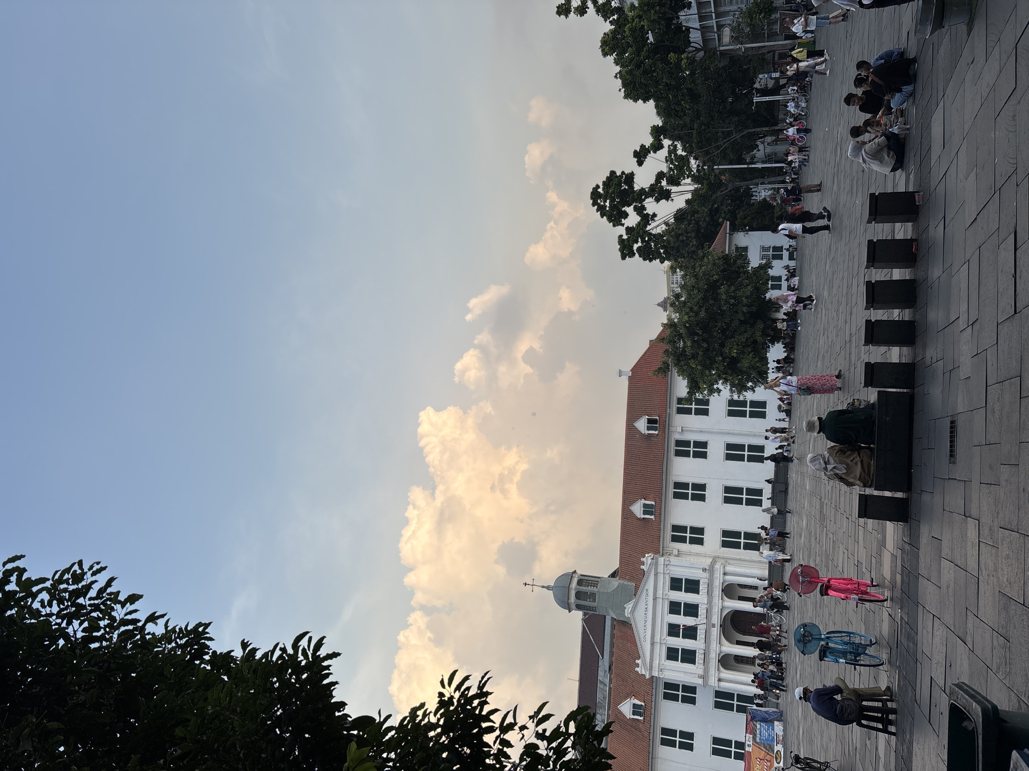 The image depicts the Fatahillah Square in the Old Town of Jakarta, West Jakarta, Indonesia. In the foreground, several people are gathered. To the left, a man wearing a white cap and blue shirt sits on a stool beside a blue bicycle. Next to him is a pink bicycle. Further to the right, two individuals, one in a light brown hijab and the other in a dark green hijab, are seated on a dark bench. A group of people are also seen sitting on the ground in the middle right of the frame, engaged in conversation. In the background, the historic white building, the Jakarta History Museum (formerly the City Hall), stands prominently with its red tiled roof. People are seen walking and congregating in front of the museum. Trees are interspersed throughout the scene, with a large tree branch in the upper left corner partially obscuring the sky. The sky is partly cloudy, with large, soft clouds illuminated by sunlight.