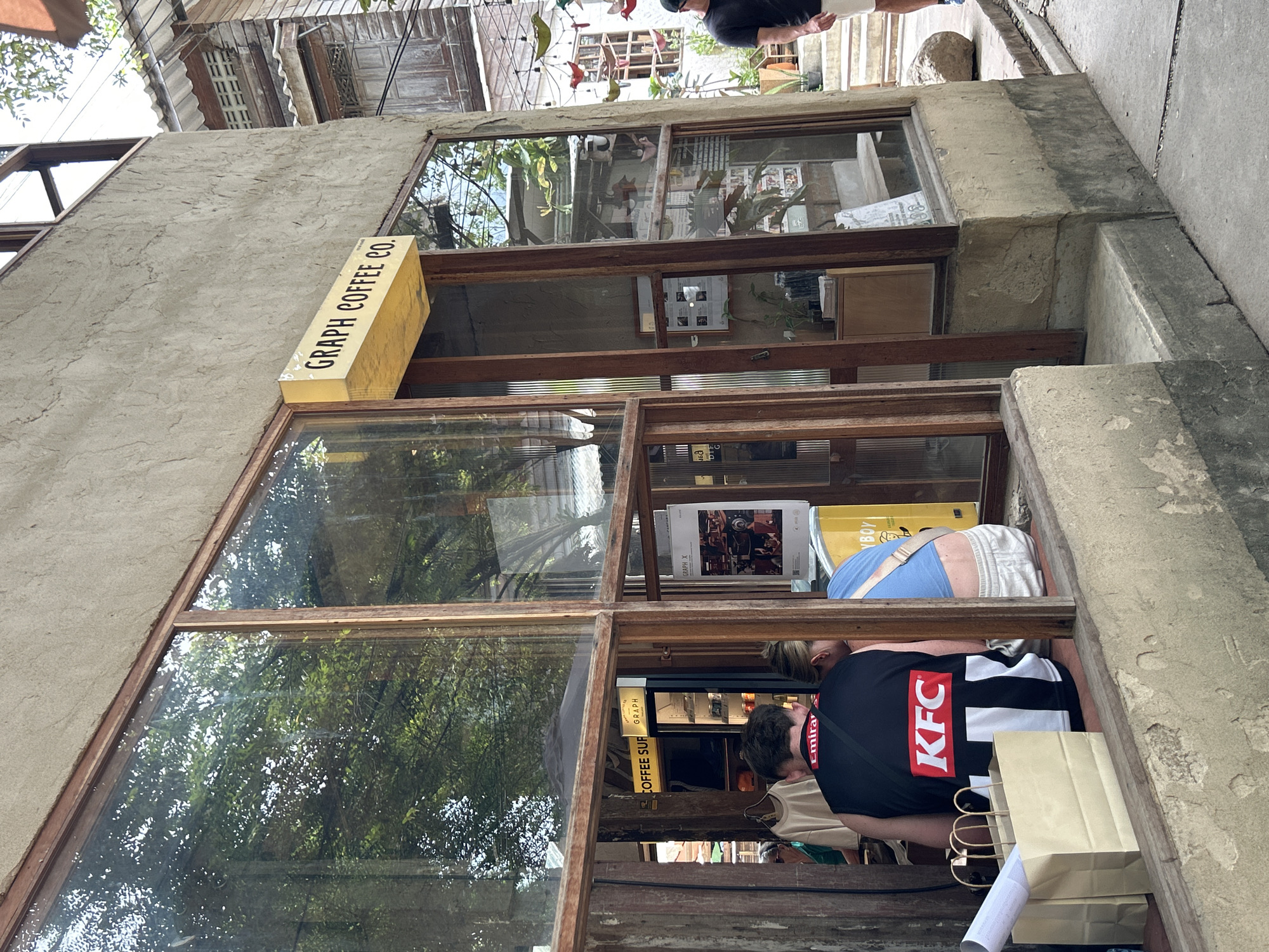The image shows the exterior of a cafe called "GRAPH COFFEE CO." in Ban Sai Kham, Thailand. The cafe features large wooden-framed windows and a rough concrete exterior. Inside, through the windows, a few people are visible. One person wears a black vest with "KFC" prominently displayed on the back. Another person, to their right, wears a blue top. In front of them, on a ledge outside the window, are two tan paper shopping bags. The cafe has a yellow sign with "GRAPH COFFEE CO." written on it. The surroundings suggest a street or alleyway with more buildings and greenery visible.