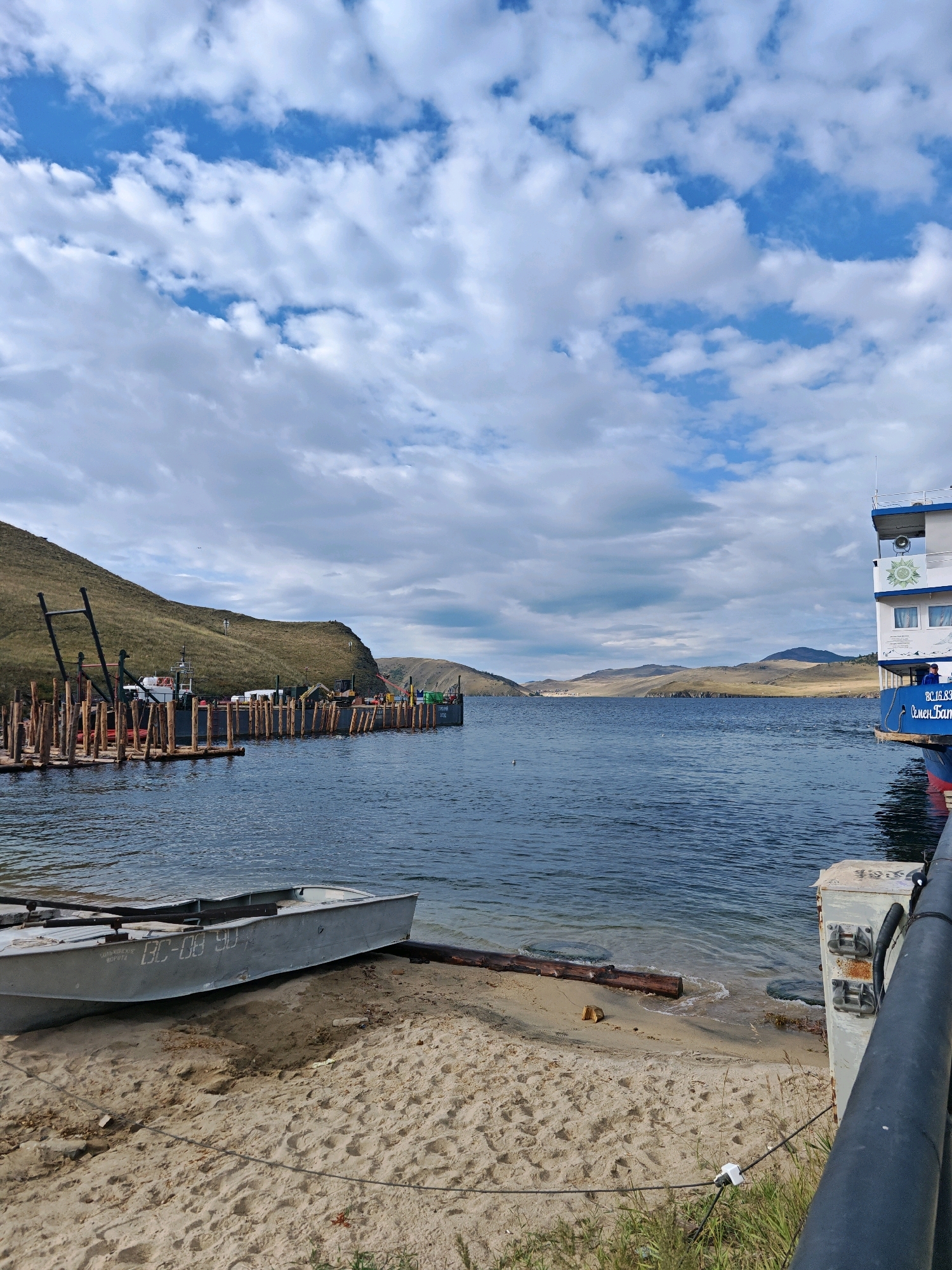The image depicts a waterfront scene in Khuzhir, Russia, characterized by a calm body of water, sandy shore, and hilly landscape. In the foreground, a small, gray metal boat with the registration "BC-08 90" is partially beached on the sand. To the right, a dark railing runs along the edge of what appears to be a pier or dock, with a white utility box mounted on it. In the middle ground, a large barge equipped with construction materials and heavy machinery, including an excavator, is moored to a wooden pier. Further in the background, a large white and blue passenger boat is visible, docked on the right side of the frame. The water is a deep blue, with gentle ripples on the surface. The shoreline is lined with sparse vegetation and a few logs. The landscape rises into rolling hills covered in dry grass, with a few scattered trees. The sky is partly cloudy, with patches of blue sky interspersed with white and gray clouds. The overall impression is of a working port or a scenic natural harbor.