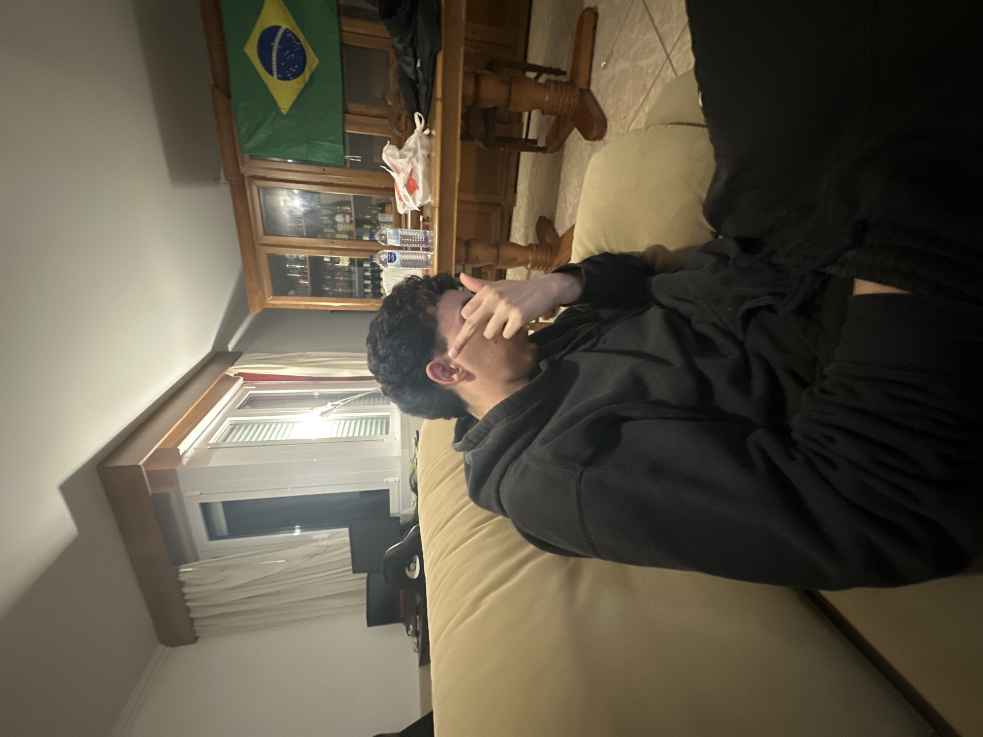A person with dark, curly hair lies on a light-colored sofa, covering their eyes with a hand. They are wearing a dark hooded sweatshirt and dark pants. The room contains a wooden cabinet displaying bottles and glassware, topped with a Brazilian flag. A wooden table with decorative legs stands near the cabinet. A window with white blinds is visible in the background, adjacent to a dark-colored object, possibly a television or monitor. The location is likely indoors, potentially in a home. I cannot confirm this is Bragança, Portugal.