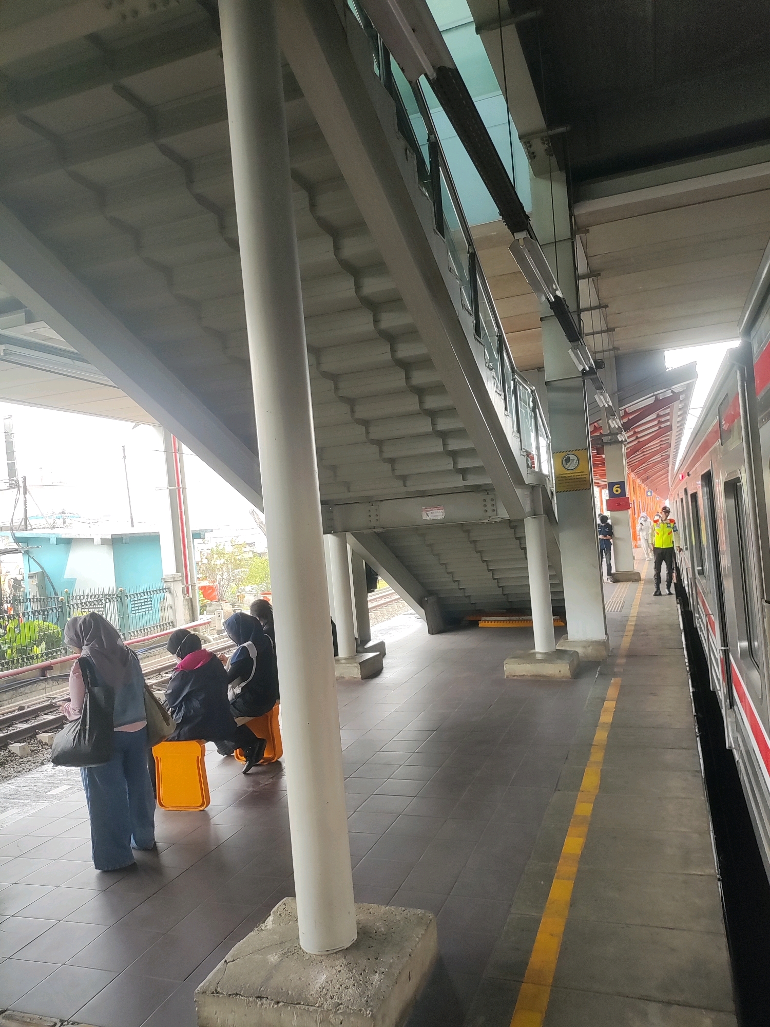 At a train station in Bekasi, Indonesia, a person wearing a bright yellow vest and a hard hat stands on the platform next to a train. The platform has a yellow line marking the safety boundary. Several other people, some wearing hijabs, are seated on bright orange stools near the tracks. Overhead, a covered walkway with visible stairs and support beams shades the platform. The overall environment suggests a functional transportation hub.