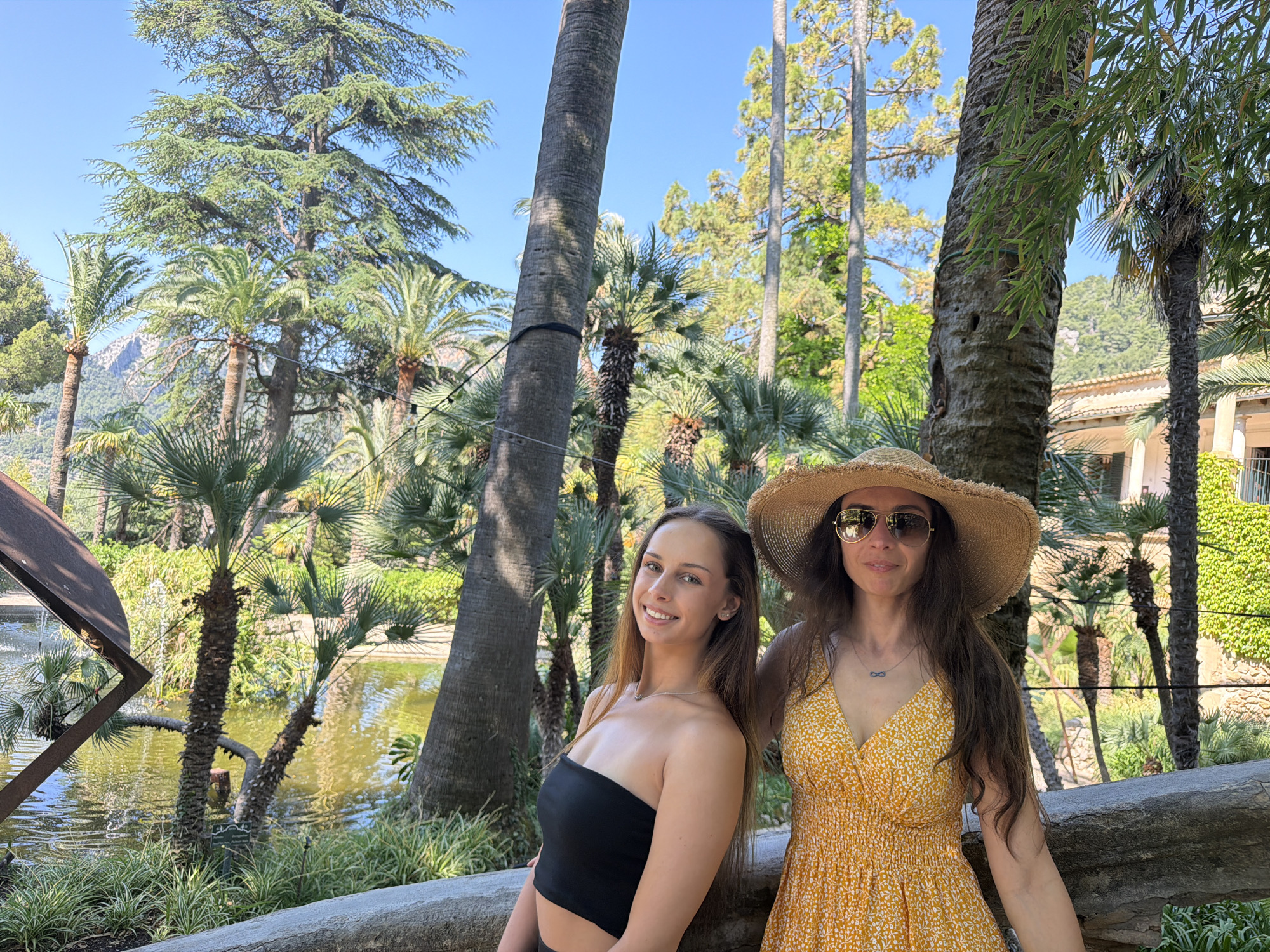 The image captures two women in a natural, outdoor setting with tropical vegetation. The woman on the left is wearing a black strapless top, while the woman on the right is wearing a yellow floral dress and a wide-brimmed straw hat with sunglasses. They are leaning against a low stone wall or barrier. In the background, there is a small lake or pond, with various palm trees and other greenery surrounding the water. The trees include palm trees and taller, broad-leafed trees. Behind them, there are structures covered with green vines, suggesting the presence of buildings. The image was taken in Bunyola, Spain.