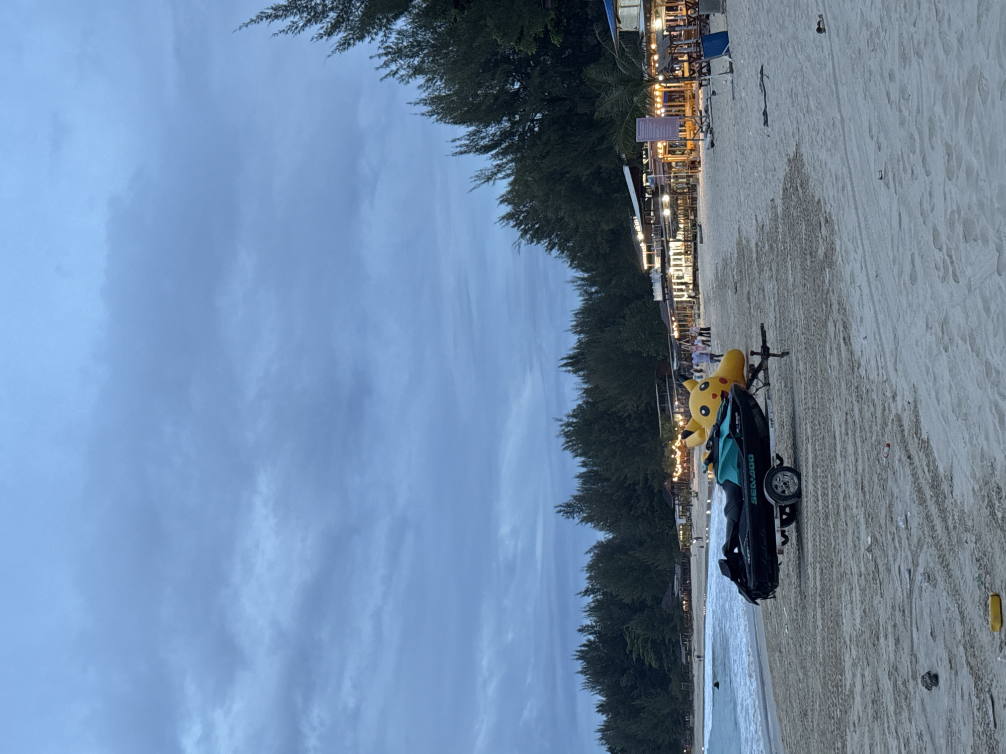 This is a photograph of a beach scene in Meunasahbale, Indonesia, taken at dusk. The sky is overcast with a muted blue and gray hue. On the sand in the foreground, a black and turquoise jet ski is loaded onto a trailer. Behind the jet ski, a large, inflatable yellow Pikachu character is visible. The beach extends towards the background where a line of buildings with illuminated lights can be seen, suggesting shops or restaurants. A dense forest of green pine trees borders the buildings and stretches across the horizon. In the distance, the ocean is visible with small waves lapping the shore, and a few people are walking along the water's edge. The sand is textured with tire tracks and scattered debris.