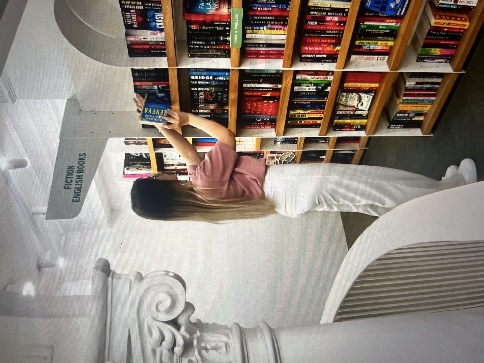 A young woman is standing in a bookstore. She is wearing white pants and a pink shirt. She is holding a book titled "GENEVA". The books are arranged on wooden shelves. A sign on the wall says "FICTION ENGLISH BOOKS". The location is Unknown, Unknown.