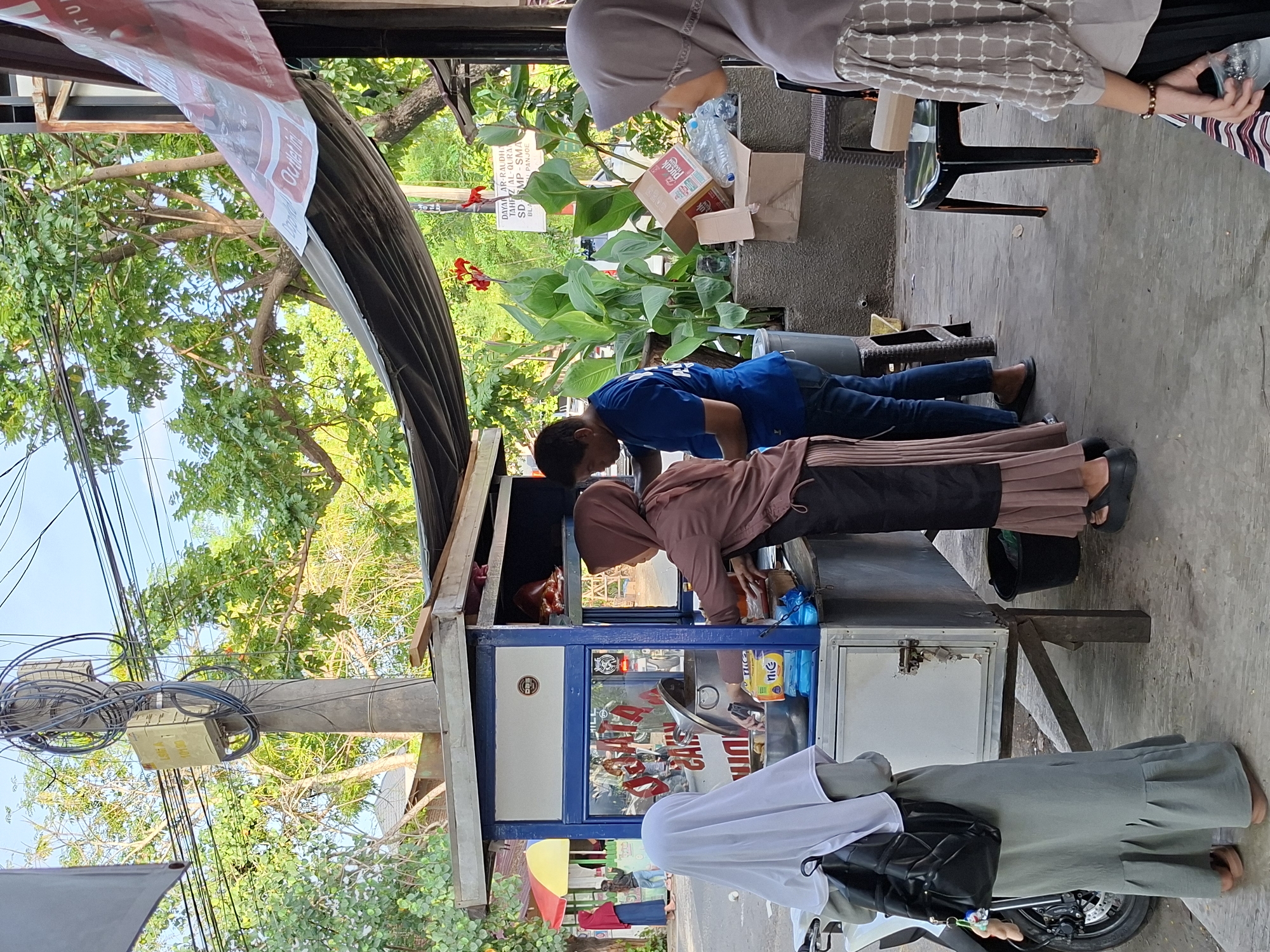 Here is a detailed description of the image:
**Scene Overview:**
The photo appears to be taken outdoors, likely on a city street in Lhokseumawe, Indonesia. The scene includes people interacting with a street food vendor. The image is oriented vertically, with a perspective that places the viewer on the sidewalk looking towards the vending stall.
**People:**
* Several individuals are present, including women wearing hijabs. One woman is wearing a gray headscarf, and another is in a brown hijab. There is a person wearing a white hijab standing next to the vehicle in front of the vending stall. Another person is wearing a blue shirt.
* The interactions observed seem to involve buying food or beverages from the vendor.
**Objects:**
* The central object is a mobile vending cart. It features a glass display case, signage, and products for sale.
* A potted plant is visible.
* Other objects include boxes, bottles, packaged goods on the cart.
* Other objects includes a black backpack, a scooter and a chair.
* The telephone pole is on the left side of the photo.
**Actions and Interactions:**
* The scene depicts commercial activity, where a vendor is selling goods to customers.
* People are standing around the vending cart, presumably engaging in transactions.
**Environment:**
* The setting is an urban environment.
* The weather appears to be daytime with bright sunlight.
* Trees and foliage are visible in the background.
* Telephone wires are strung overhead.