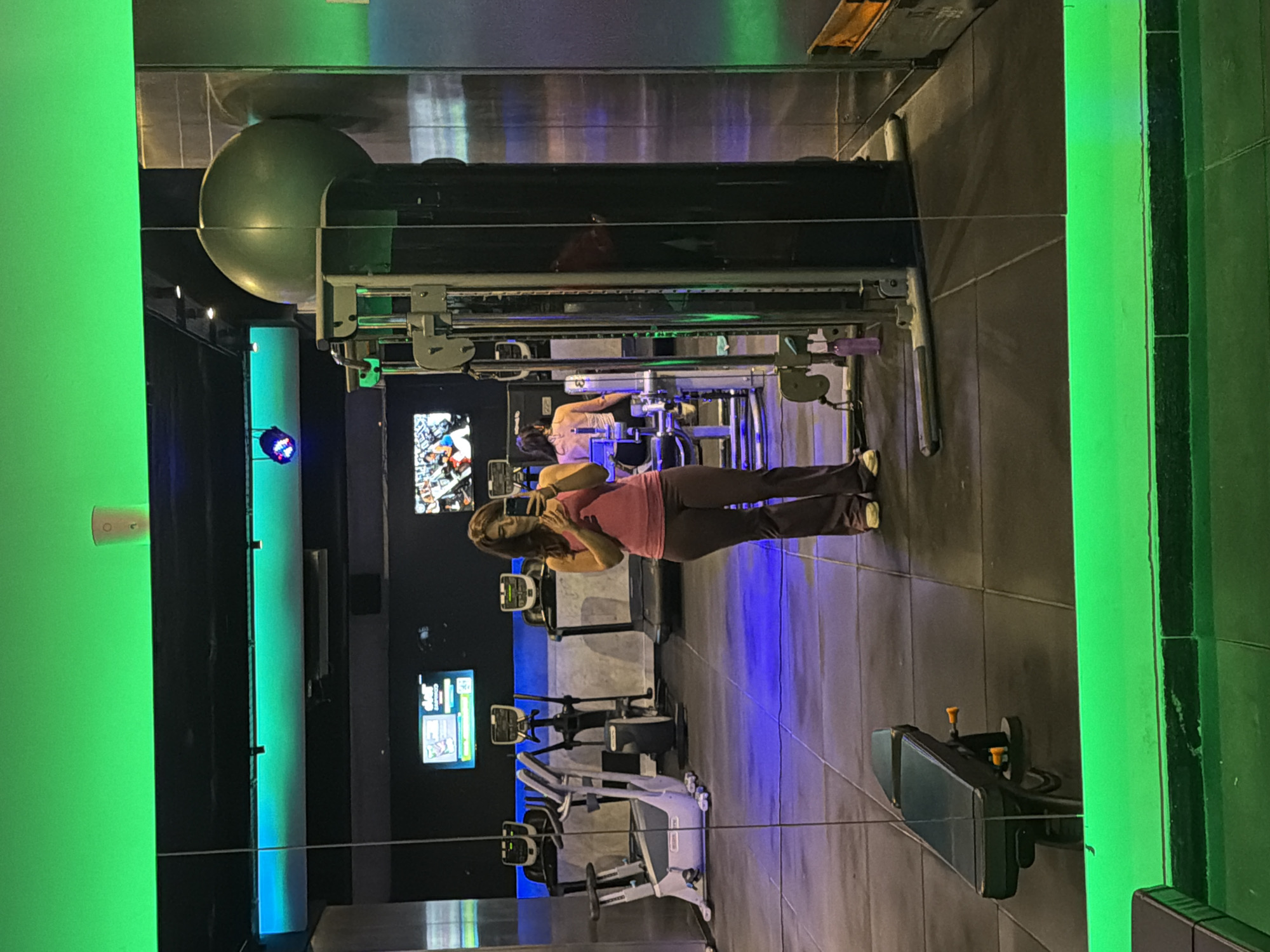 The image displays the interior of a modern gym, viewed as a reflection in a mirror or glass surface. The setting features dim ambient lighting dramatically enhanced by colorful artificial illumination.
In the reflected foreground, a woman with long, dark hair, wearing a pink top and dark pants, is captured taking a selfie with a smartphone held at her face. Her body is angled slightly, facing the reflective surface.
Behind her, within the reflection, various gym equipment is visible. A prominent black cable machine stands closer to the foreground, accompanied by a large green spherical object, likely a stability or medicine ball, positioned near its top. Further in the background, another woman appears to be using a piece of exercise equipment. Several ellipticals or exercise bikes are also discernible on the gym floor.
The gym's atmosphere is defined by bright neon green vertical lights accenting the walls and blue lights illuminating sections of the floor around the machines. Multiple digital screens are visible in the background, displaying graphics, but no specific text is clearly legible. The overall impression suggests an indoor evening or night setting due to the reliance on artificial lighting. The location context provided is Buenos Aires, Argentina.